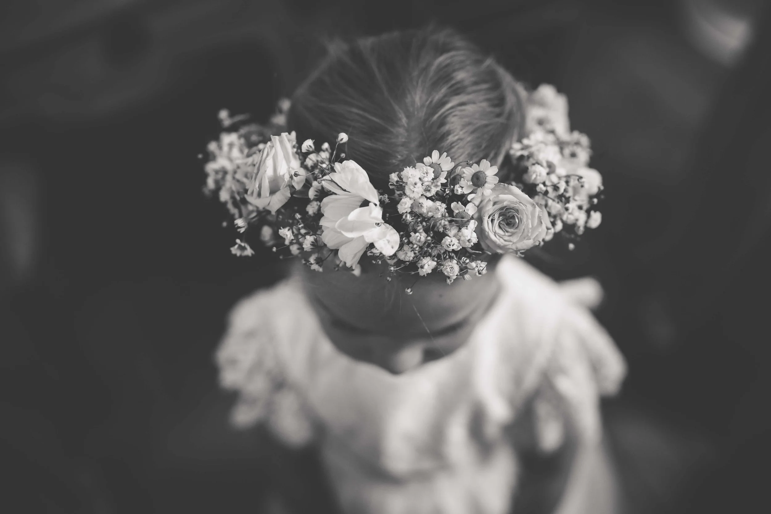 A girl wearing a flower crown of roses and smaller flowers, looking down, with a blurry background.
