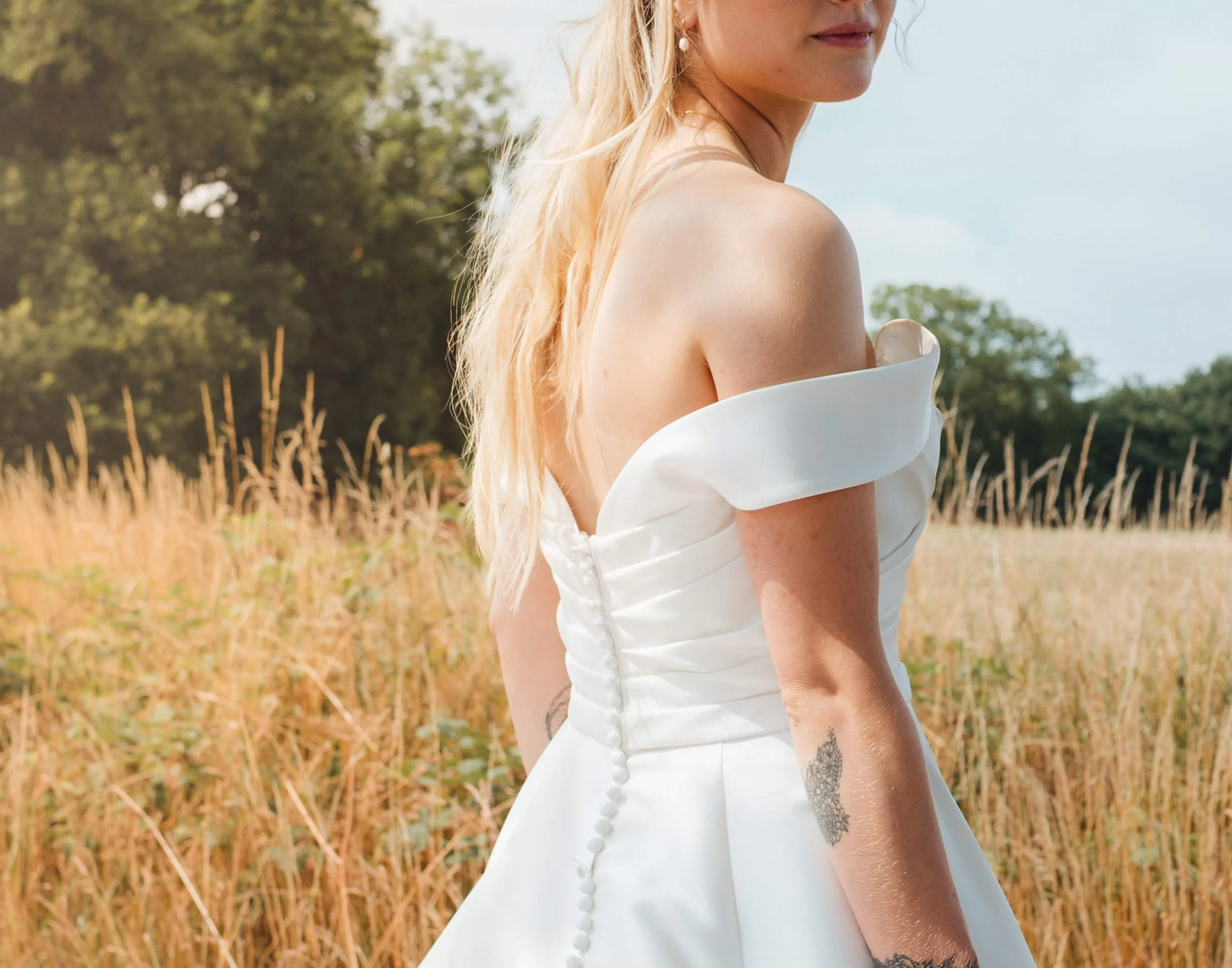 A woman in a white off-shoulder wedding dress standing in a field of tall grass with trees in the background.