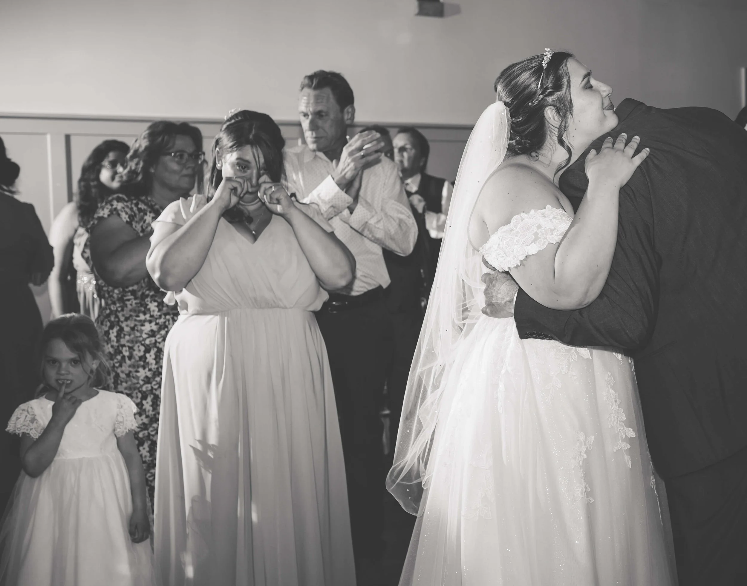 A bride and groom share a kiss at their wedding, surrounded by emotional guests including a woman wiping tears and a young girl standing in front of a woman with glasses.