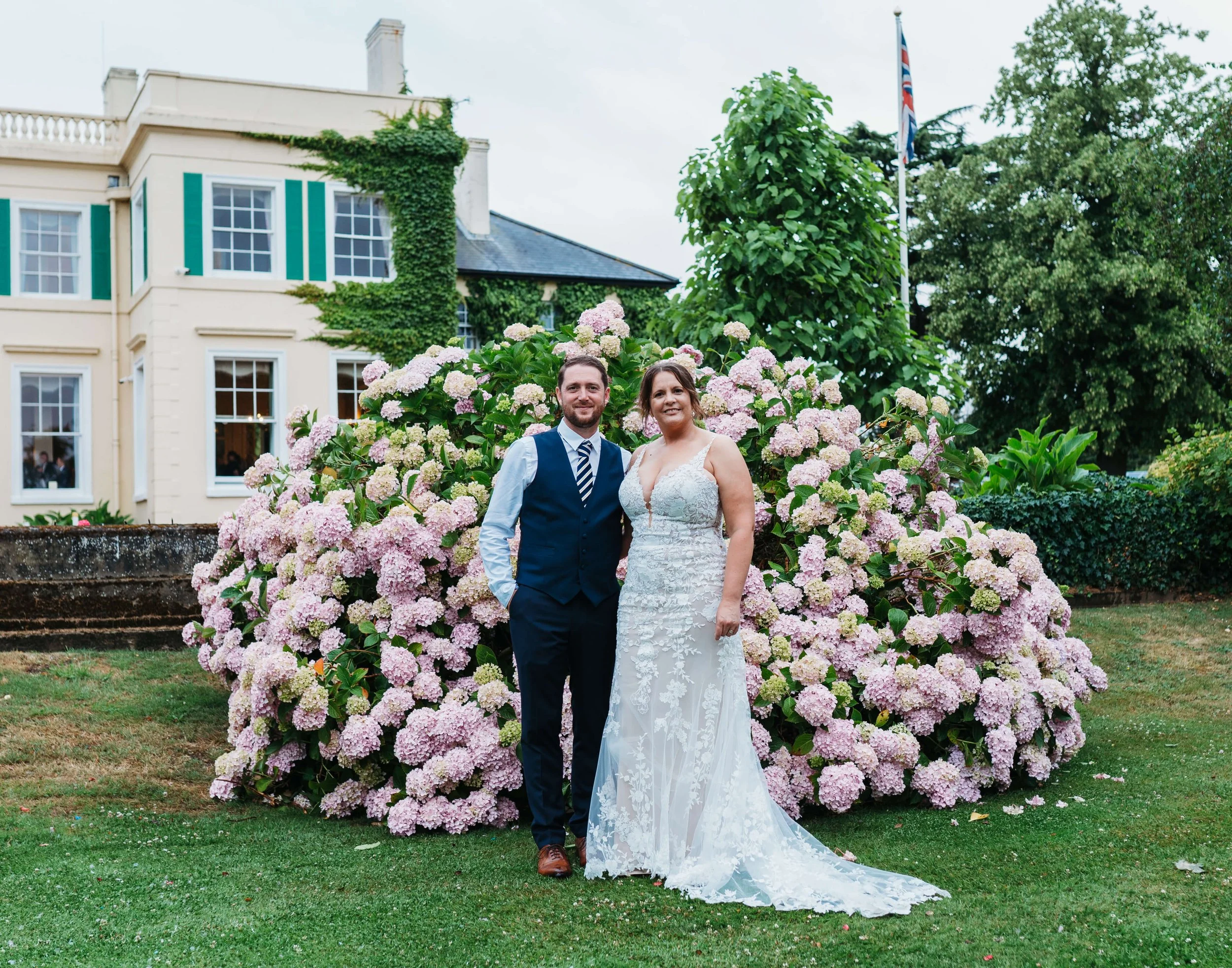 A newlywed couple standing in front of a large pink hydrangea bush outside a historic house with green shutters and ivy climbing its walls. The groom is wearing a navy vest, tie, and white shirt, and the bride is in a lace wedding gown. The backgroun
