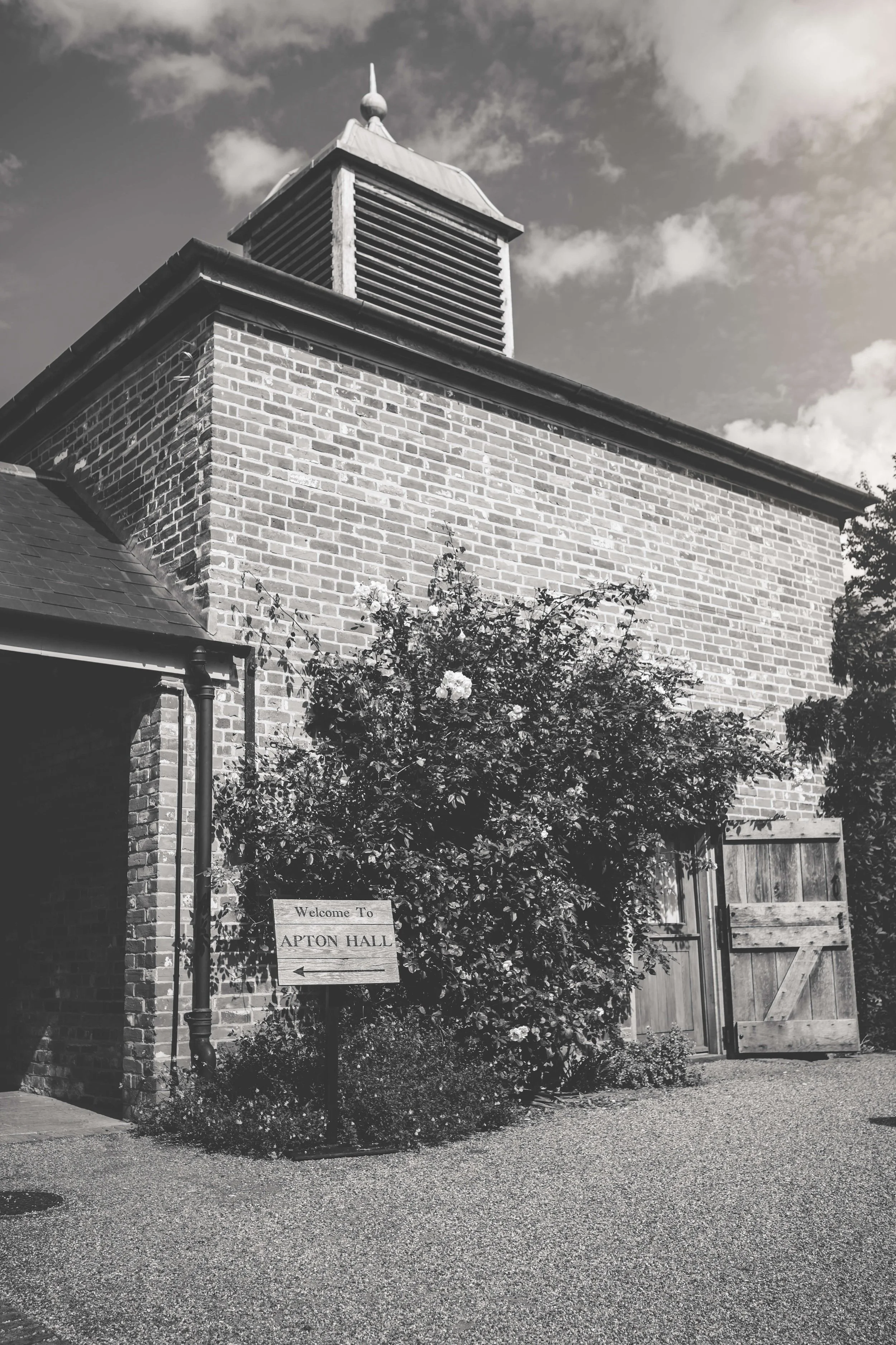Black and white photo of a brick building with a bell tower, a bush with flowers in front, a wooden gate, and a sign that reads "Welcome To APTON HALL" with an arrow pointing left.
