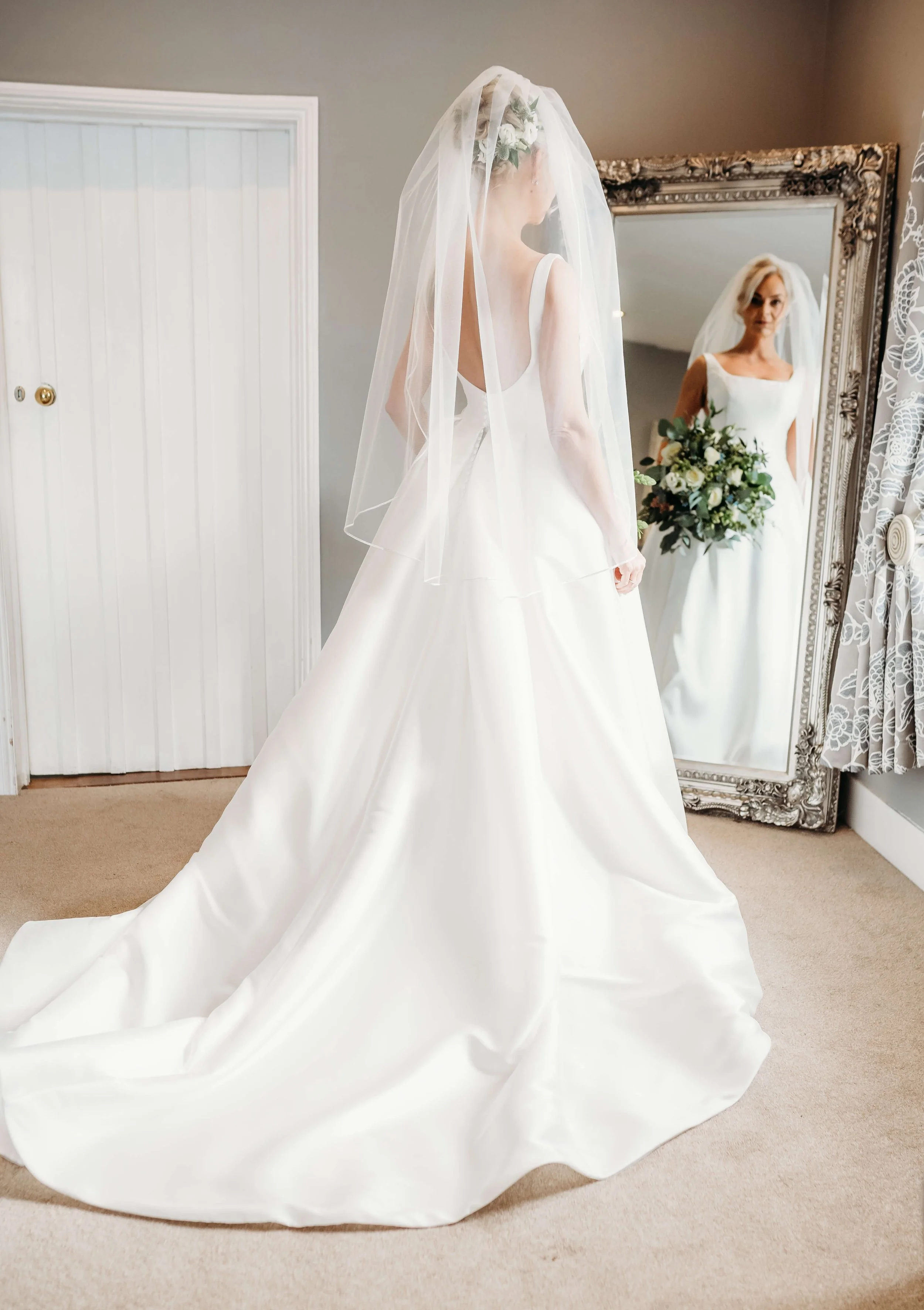 Bride in a white wedding dress with a veil looking in a mirror, holding a bouquet of flowers.
