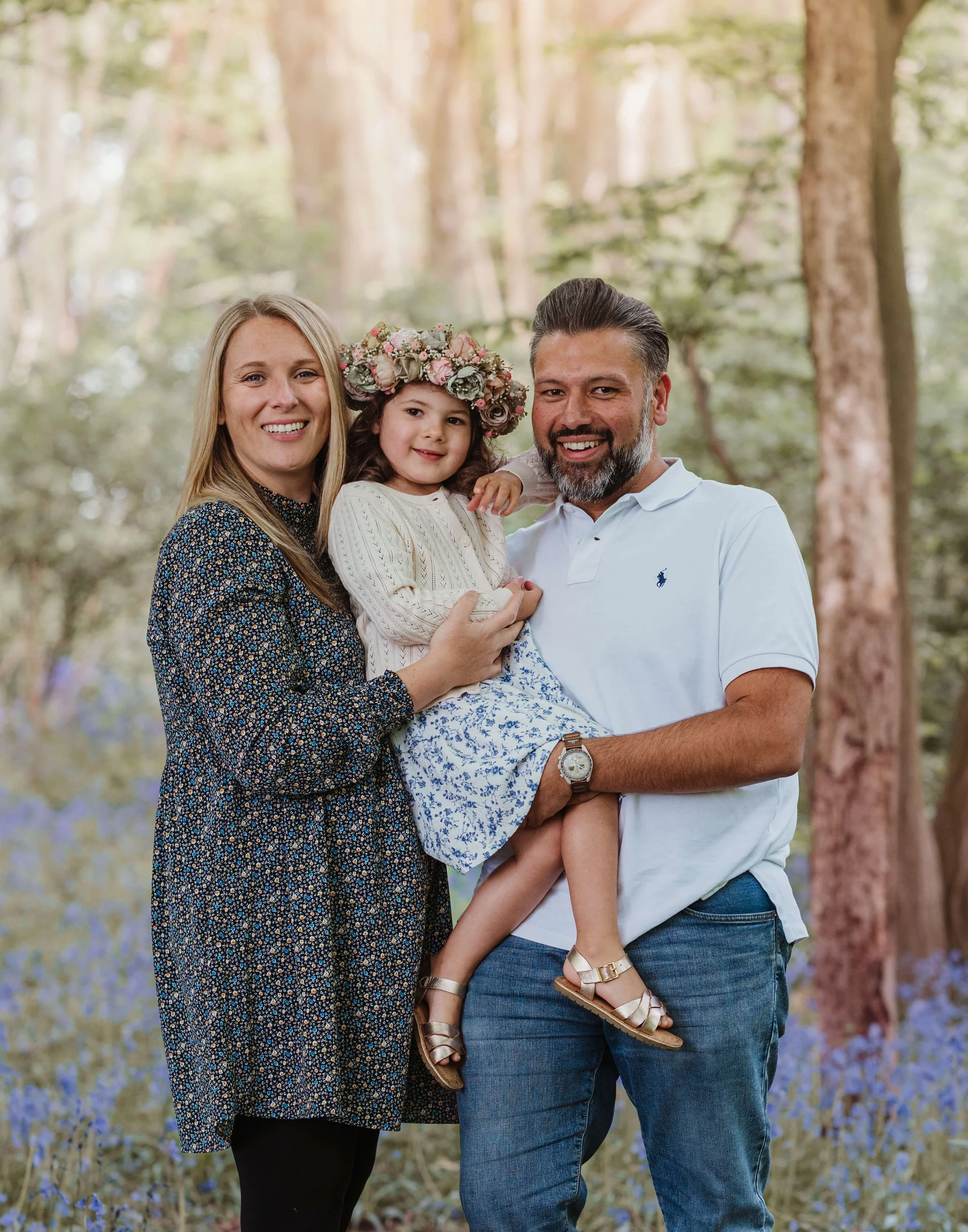 A family of three smiling in a wooded outdoor setting with purple flowers in the background. The woman has blonde hair and is wearing a floral dress, holding a young girl wearing a flower crown and a white sweater. The man has dark hair and a beard, 