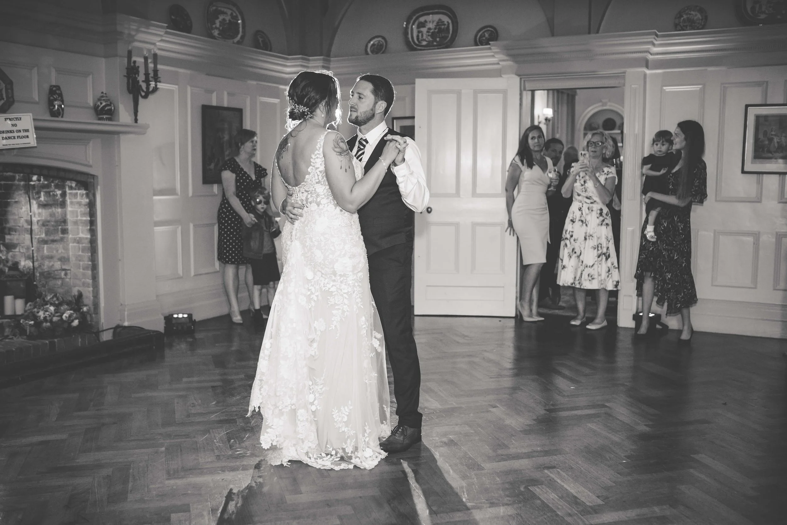 A bride and groom are dancing together in a ballroom. The bride is wearing a lace wedding dress, and the groom is in a suit with a striped tie. Several women and a child are watching from the background.