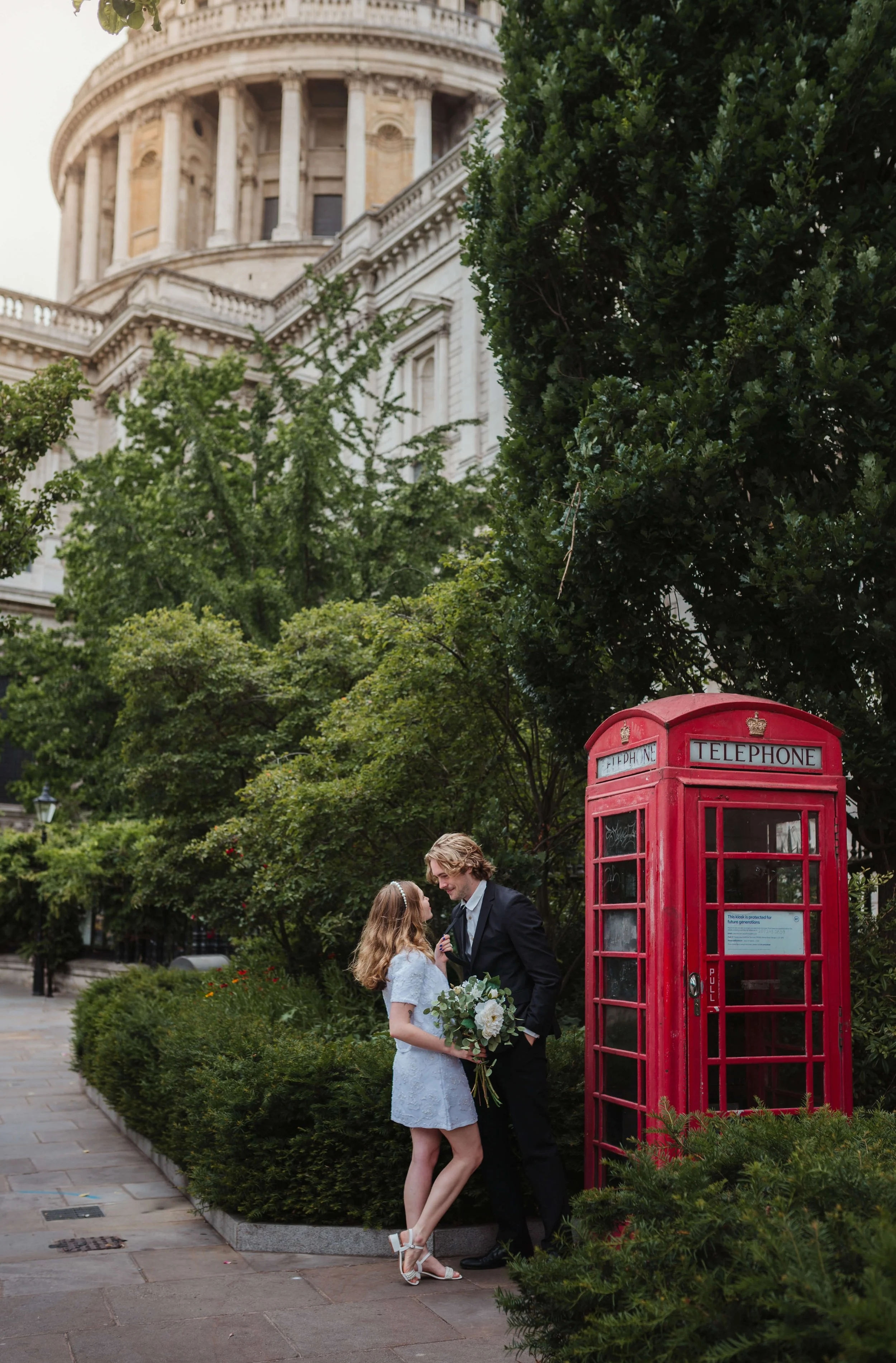 A young couple is standing next to a red British telephone booth on a sidewalk, surrounded by green trees and bushes. The woman is wearing a white dress and heels, holding a bouquet of flowers, while the man, dressed in a black suit, is leaning in to