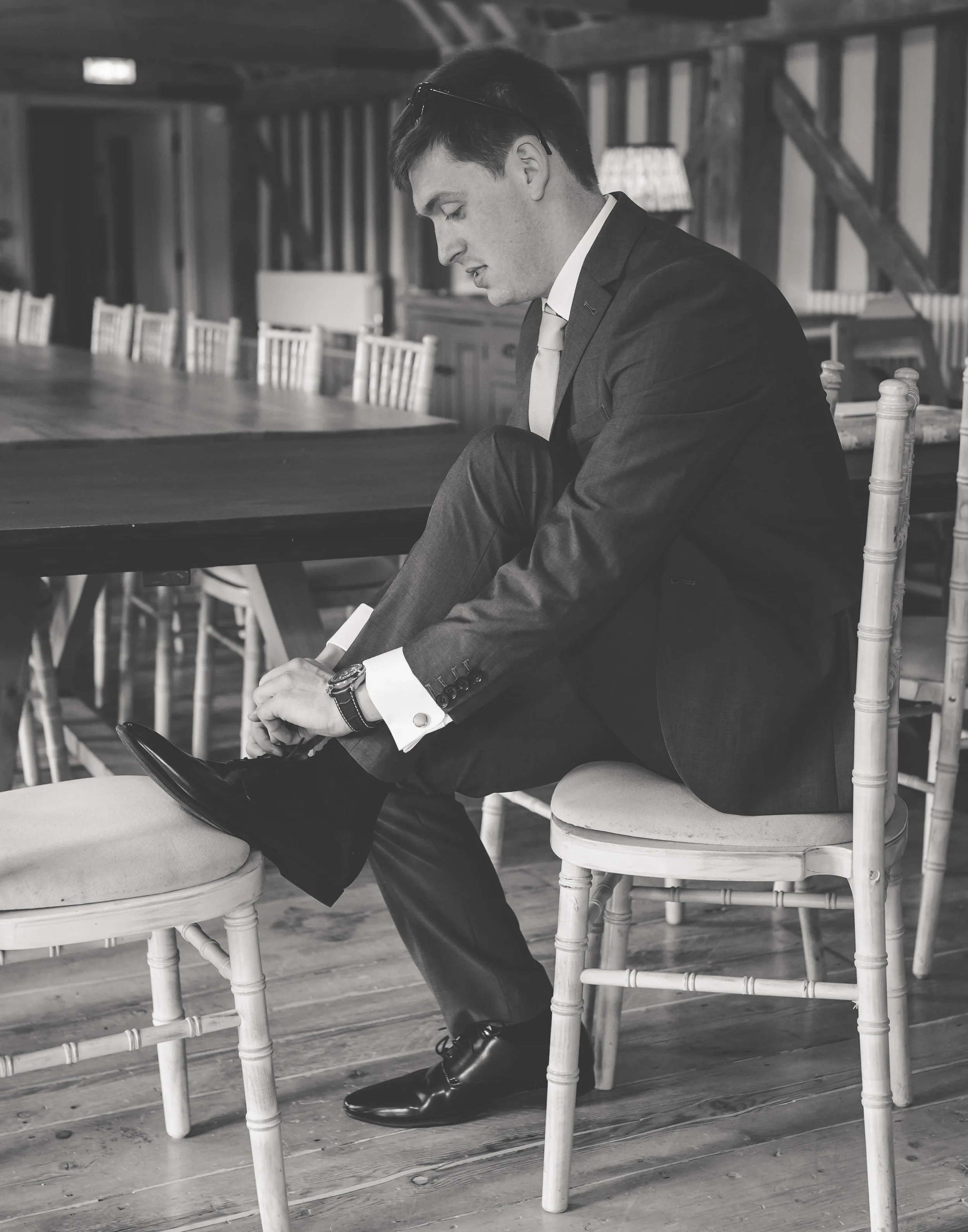 A man in a suit sitting on a chair in a rustic room, adjusting his shoe.