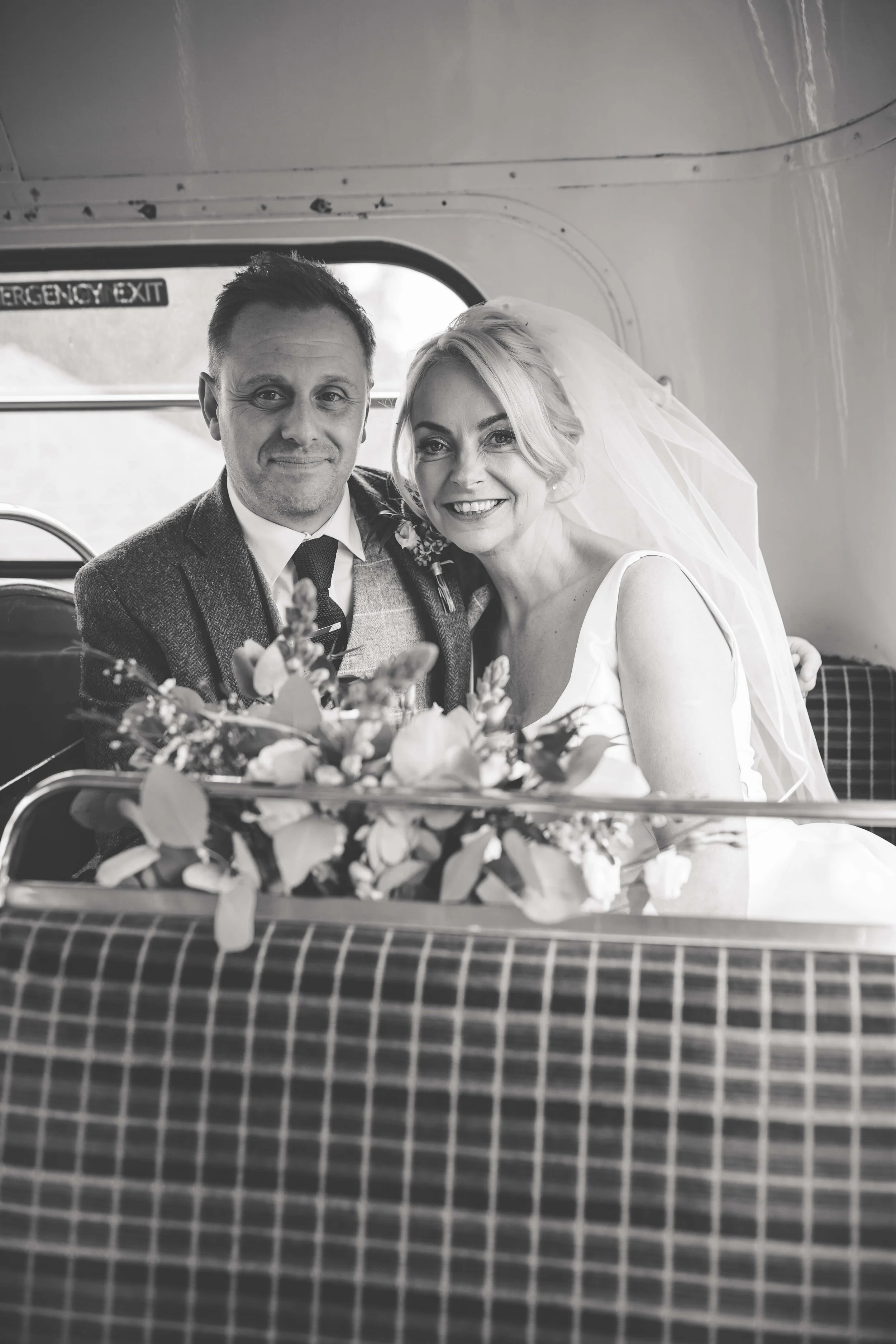 A black and white photo of a groom and bride sitting together inside a vehicle, with a bouquet of flowers in front of them. The groom is wearing a suit and tie, and the bride is wearing a wedding dress and veil, smiling at the camera.