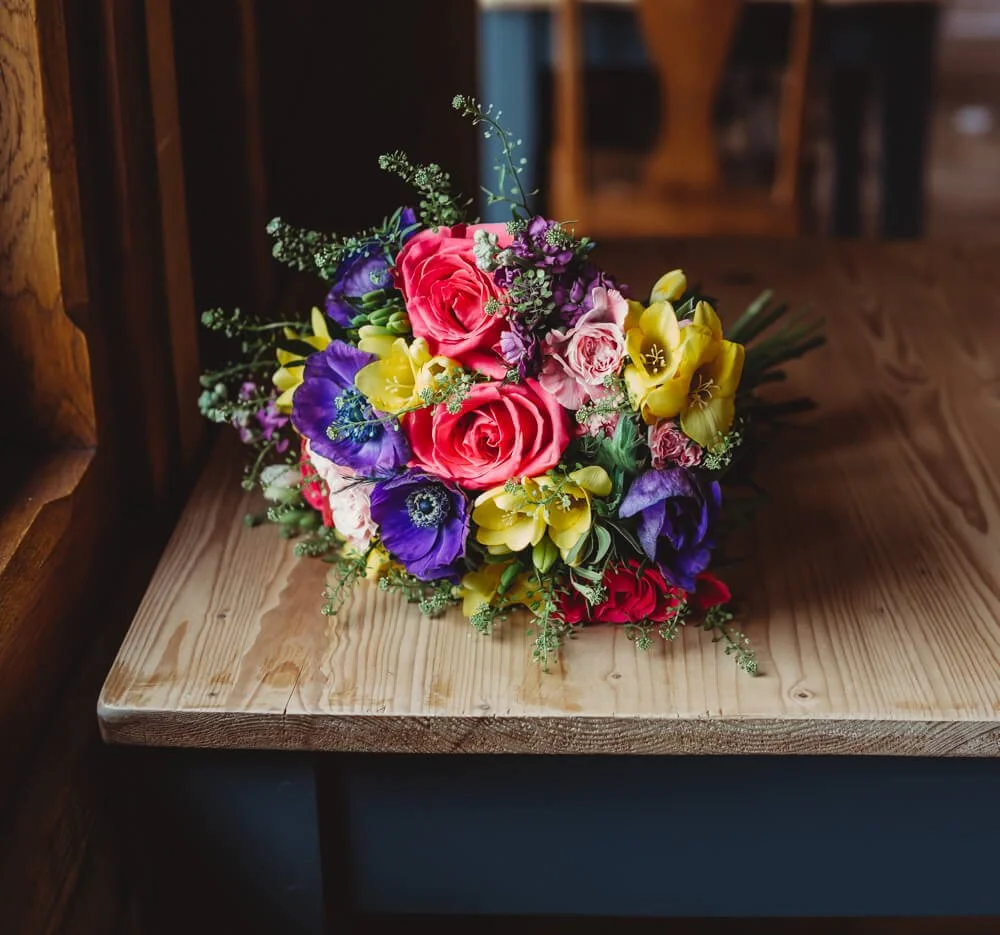 A colorful bouquet of flowers with pink, purple, yellow, and red blooms resting on a wooden surface with a dark background.