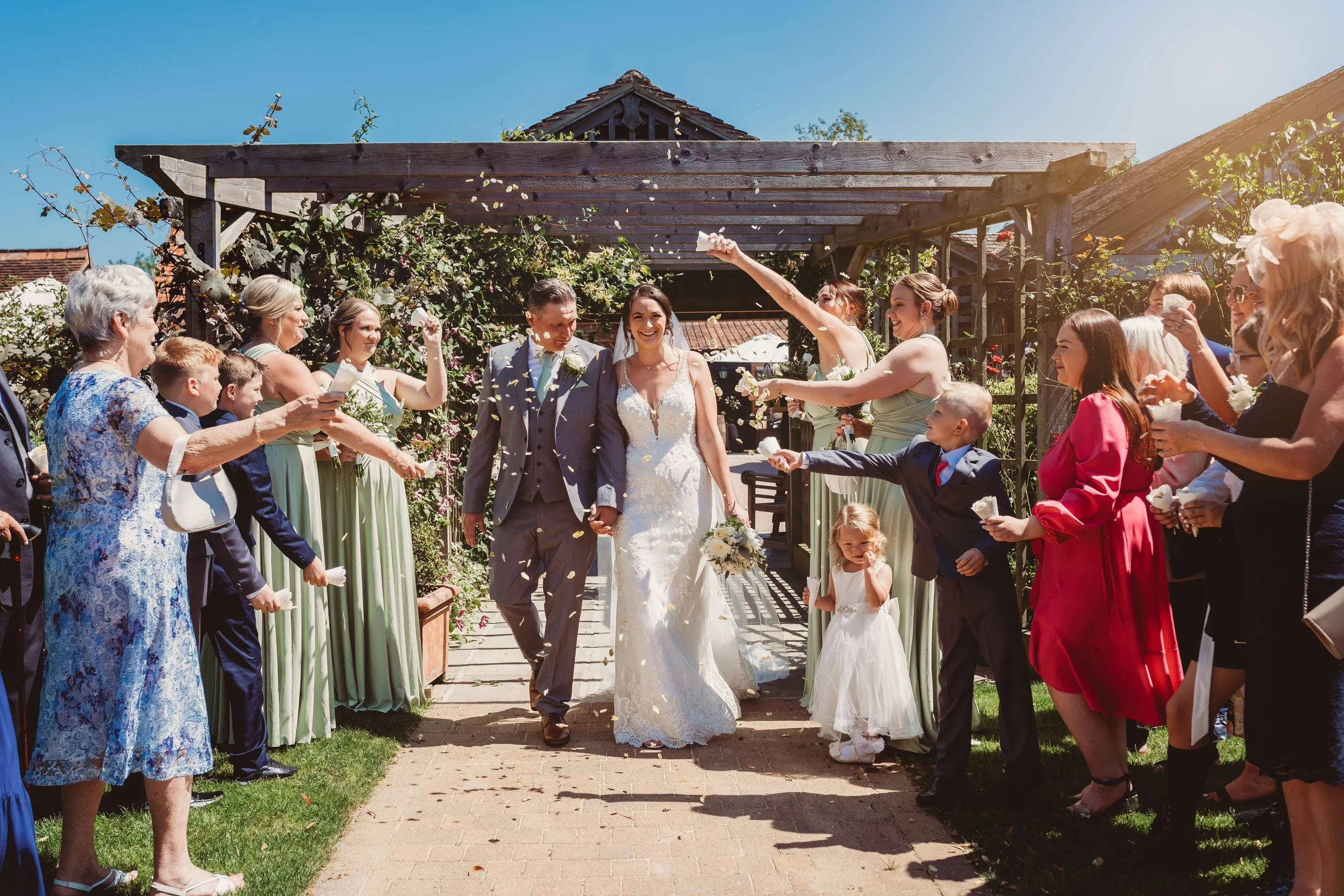 A bride and groom walking outdoors through a garden arch, surrounded by friends and family celebrating their wedding with confetti and flower petals on a sunny day.