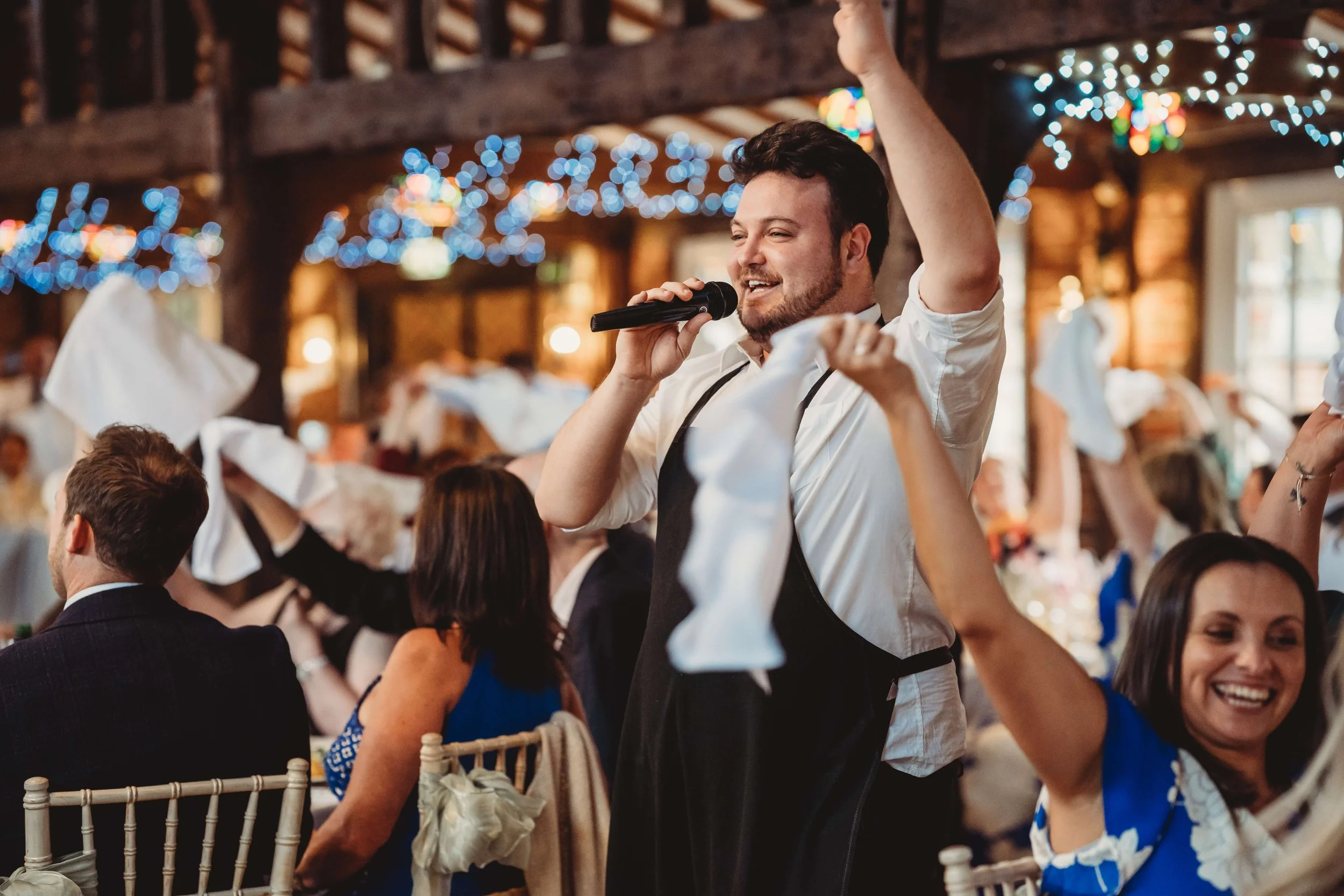 Man holding microphone and raising his arm at a lively celebration or wedding reception, surrounded by guests clapping and smiling in a decorated venue with colorful string lights.