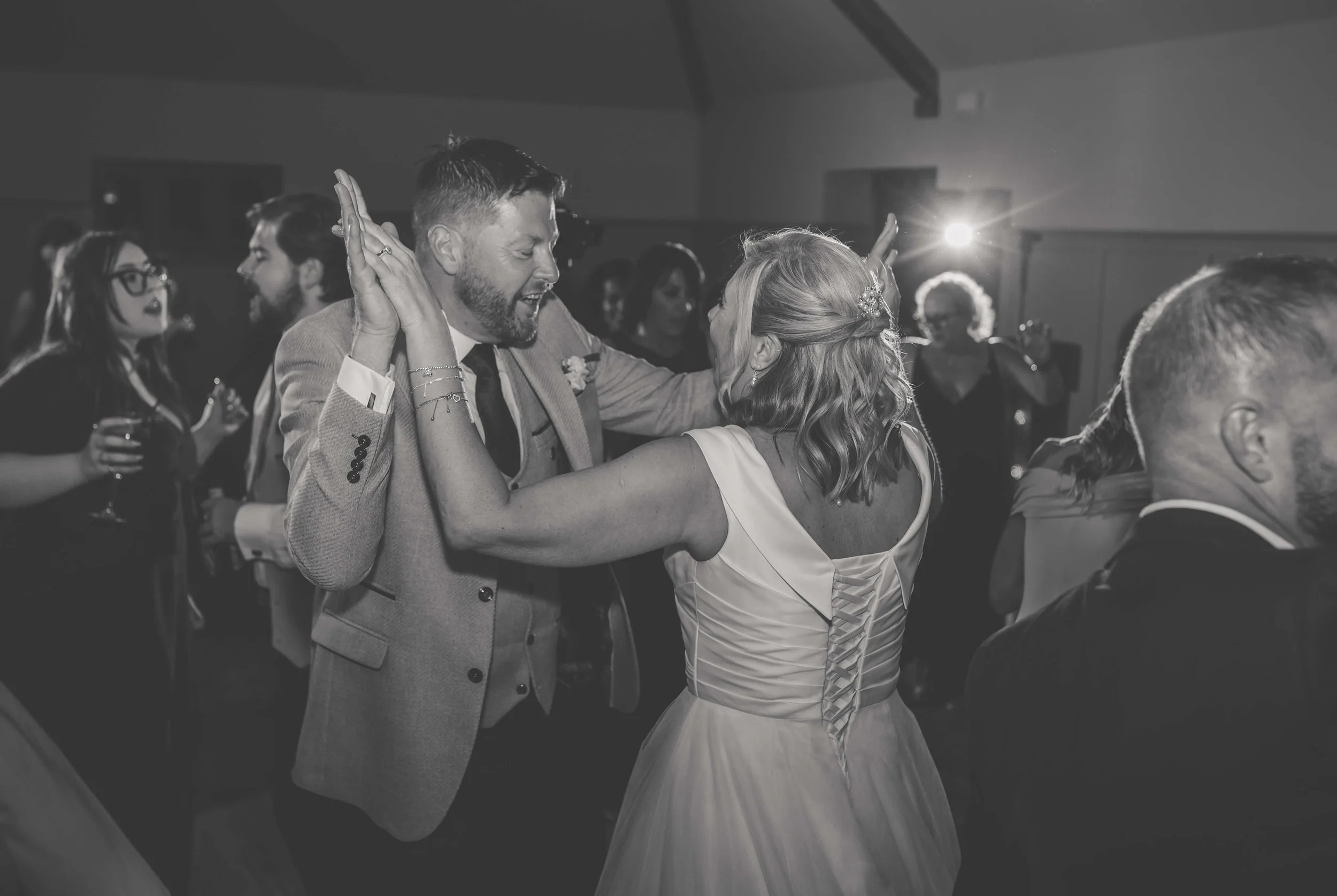 Black and white photo of a couple dancing at a wedding reception, surrounded by other guests, with one woman wearing a white wedding dress and a man in a suit.