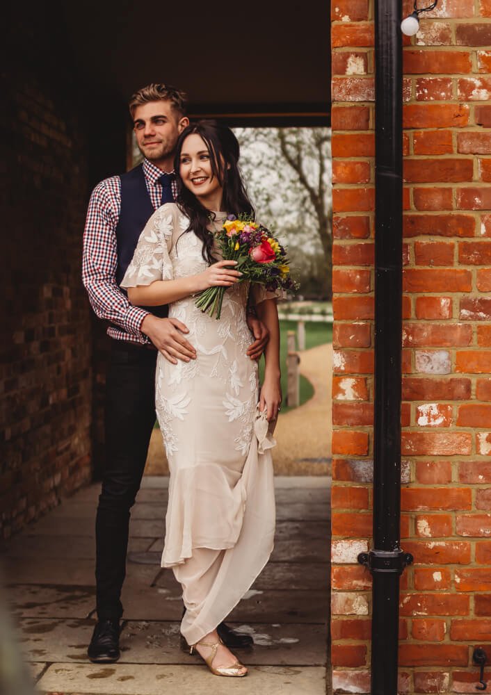 A happy couple in wedding attire standing closely together outside near a brick wall, with a woman holding a bouquet of flowers and a man embracing her from behind.