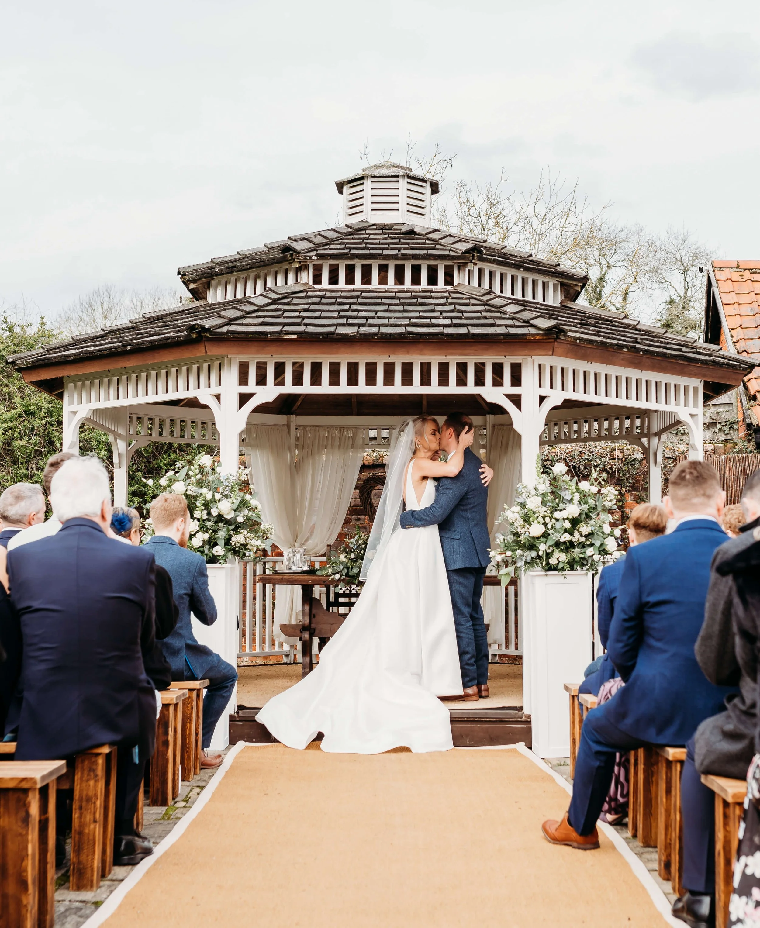 A bride and groom share a kiss during their outdoor wedding ceremony under a white gazebo decorated with flowers, with guests seated on either side.