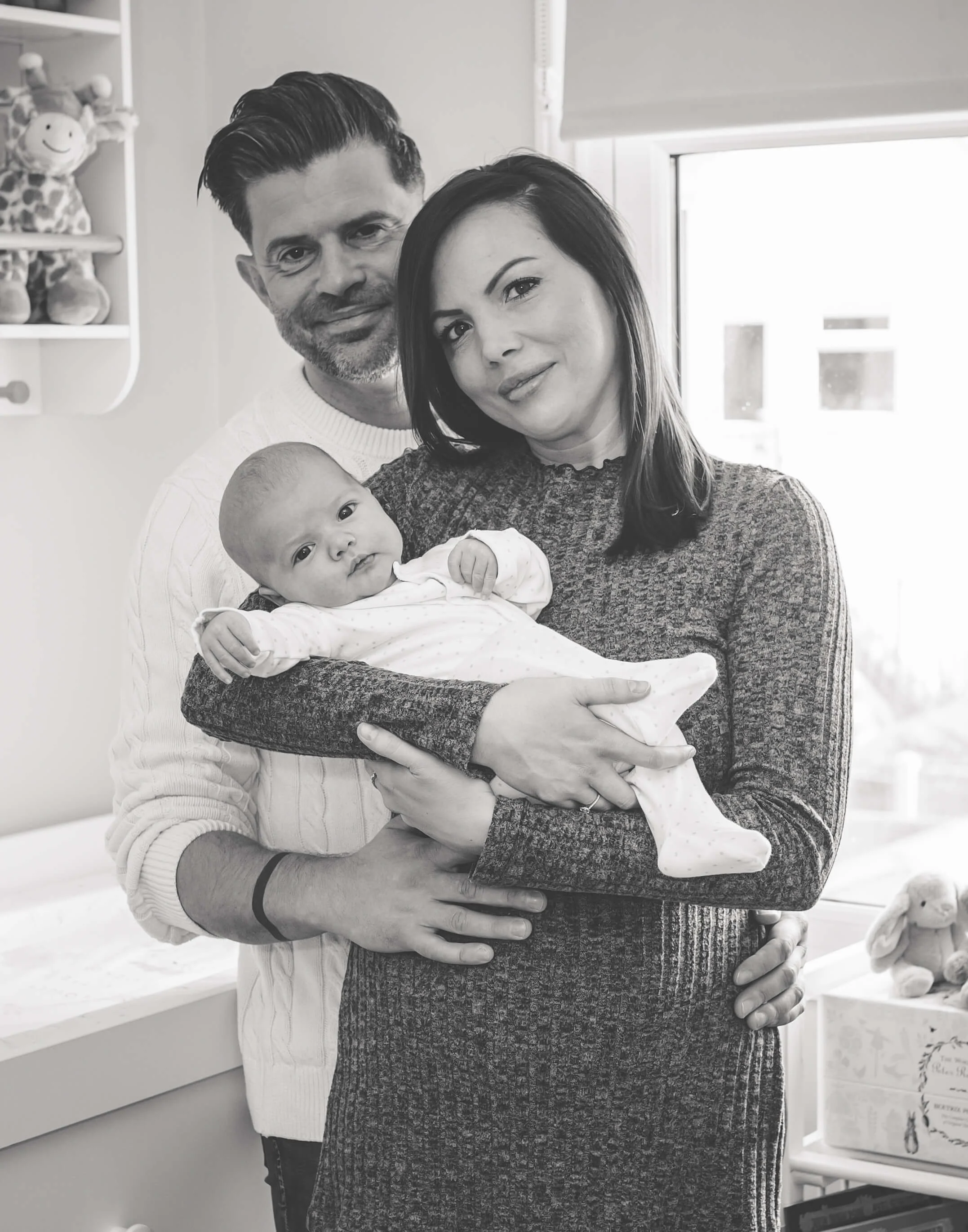 Family of three, including a woman holding a baby, with a man standing behind them, in a cozy room with toys and decorations.