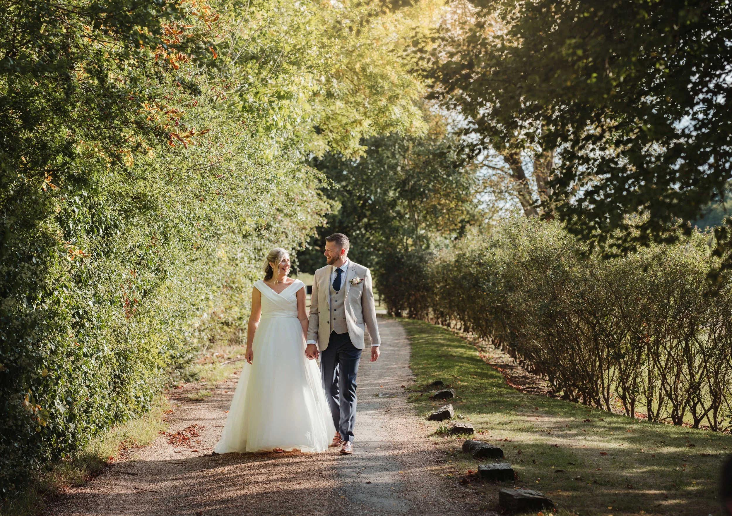 A newlywed couple walking hand in hand through a lush, green park pathway surrounded by trees and bushes on a sunny day.
