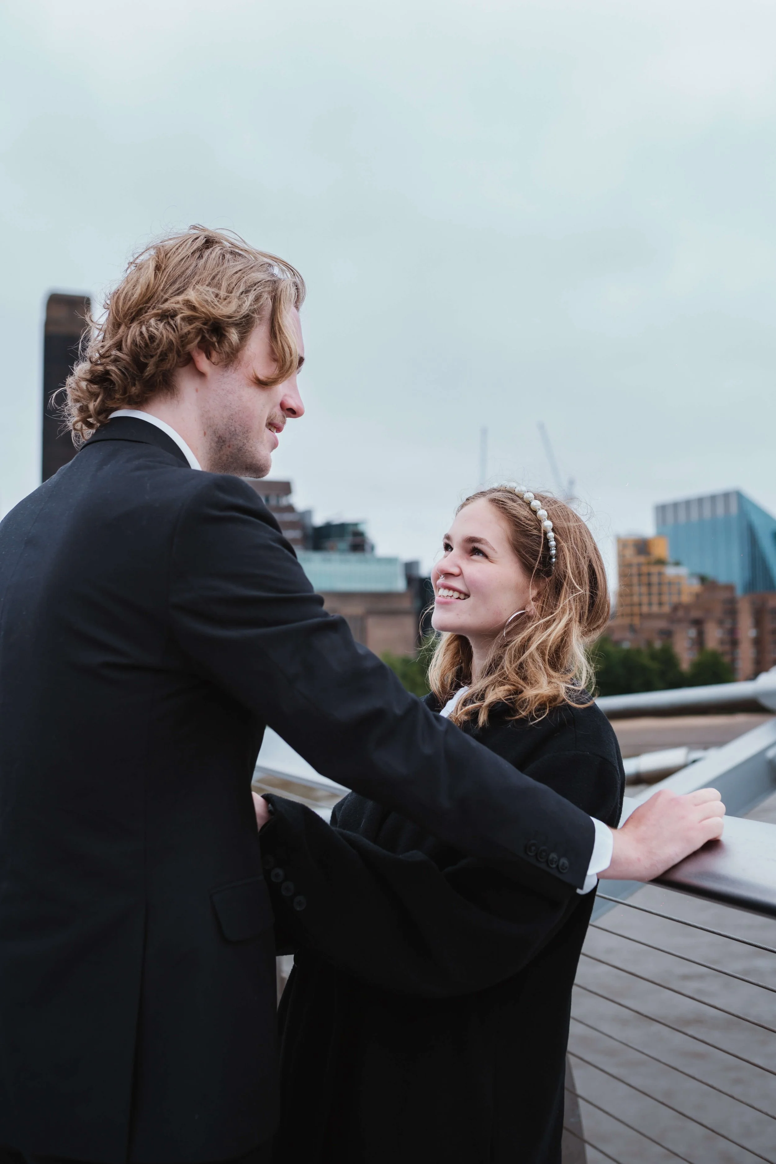 A man and woman on a rooftop, gazing at each other, city skyline in the background.
