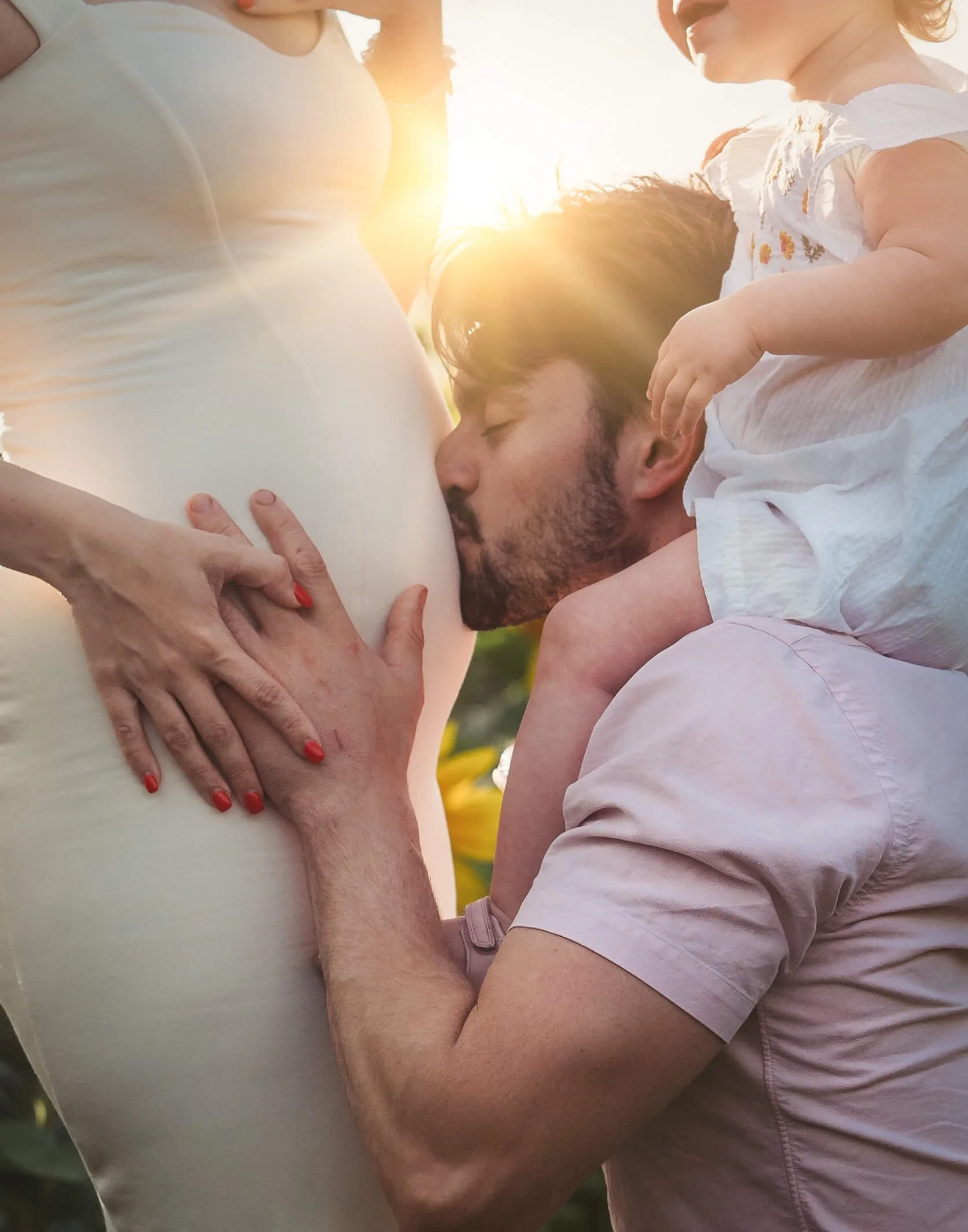dad kisses mums pregnant belly with daughter on his shoulders with a beautiful sun flare shining through 