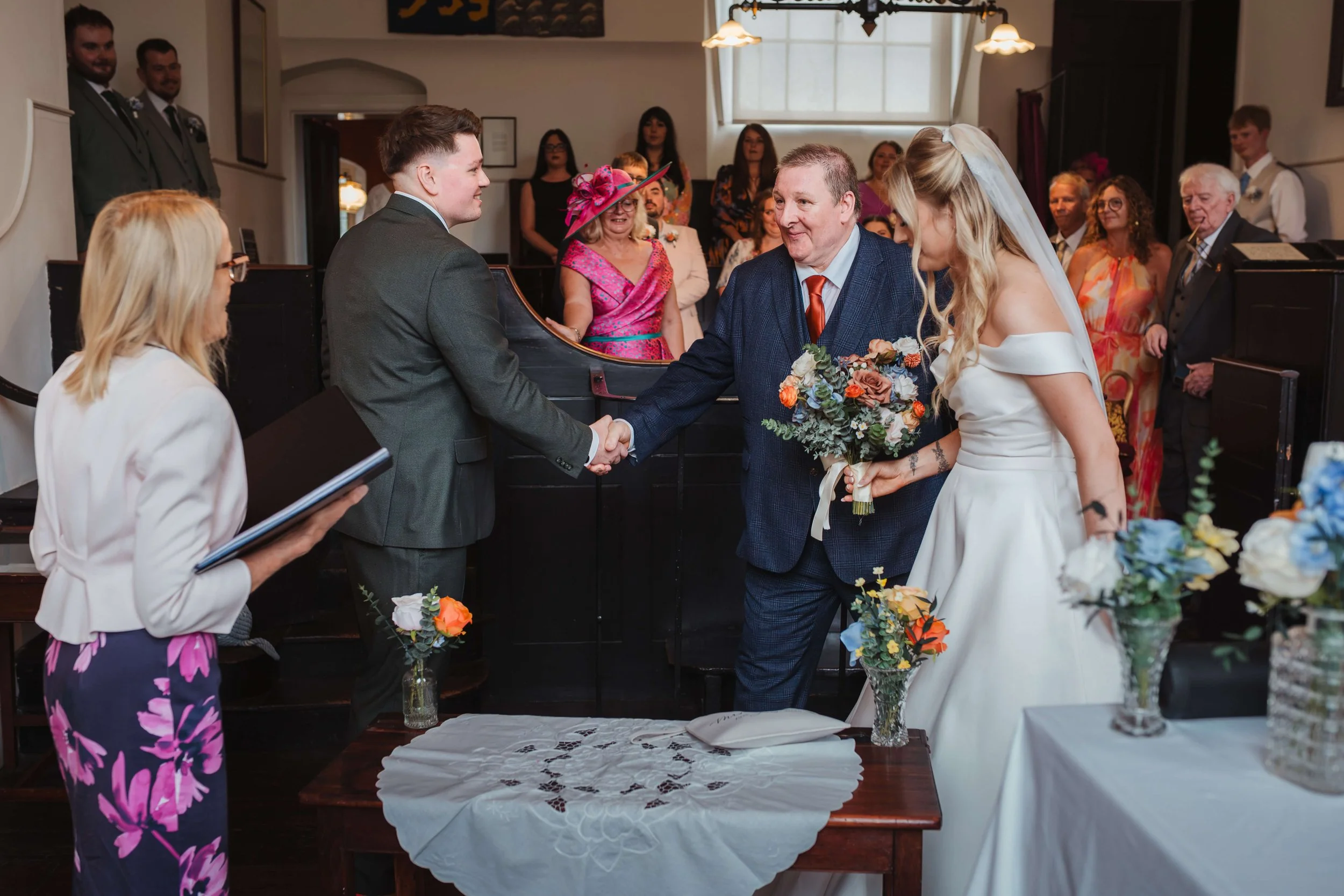father of bride and groom shake hands as father gives bride away at ceremony with emotional guests 