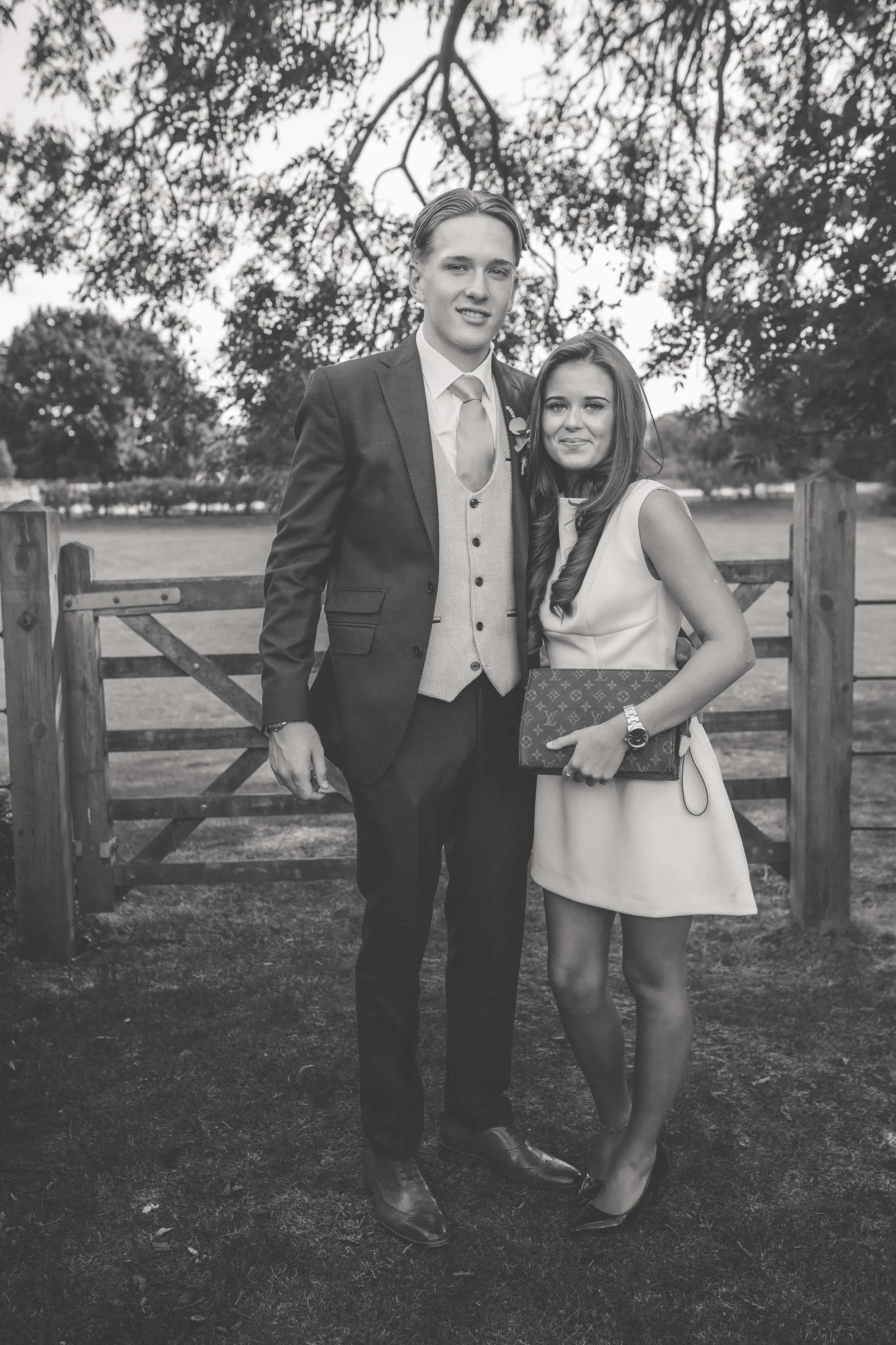 A young man and woman dressed in formal attire standing outdoors near a wooden fence, with trees in the background.
