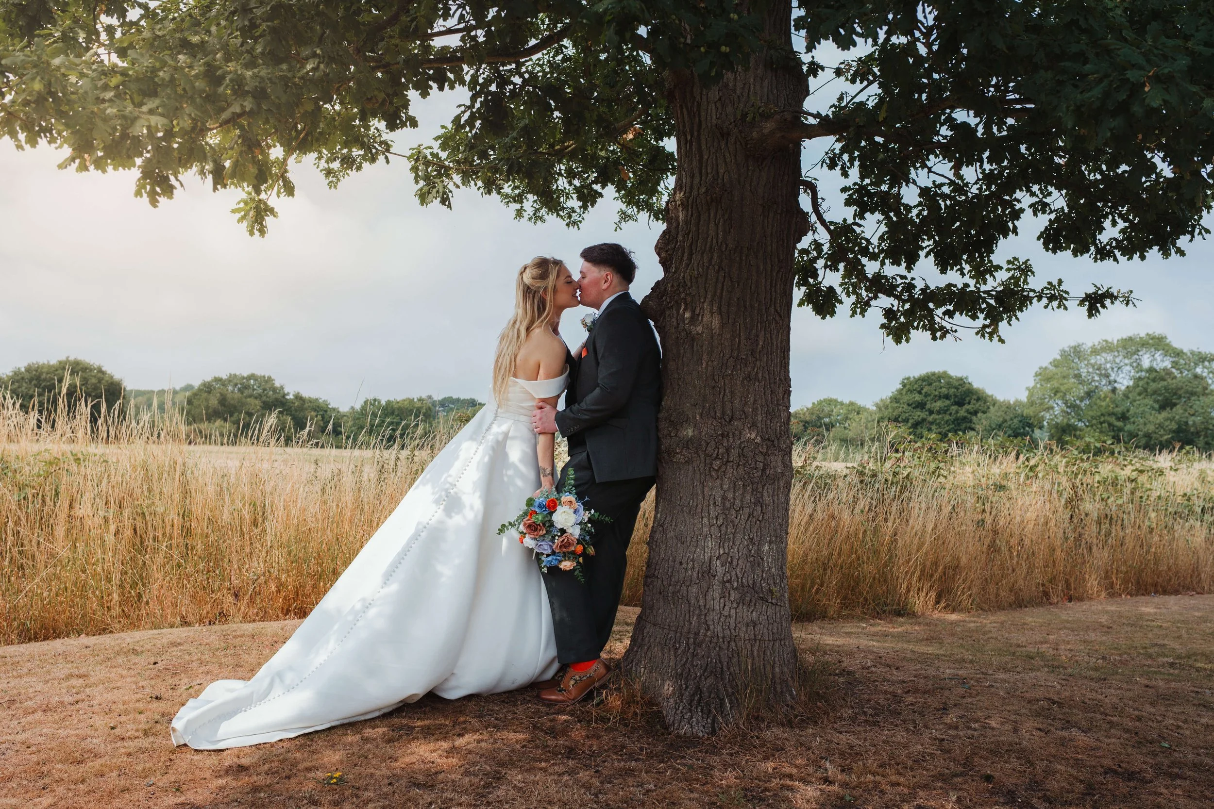 Wedding couple kissing under a large tree in a field, bride in white gown holding bouquet, groom in black tuxedo, outdoors during daytime.