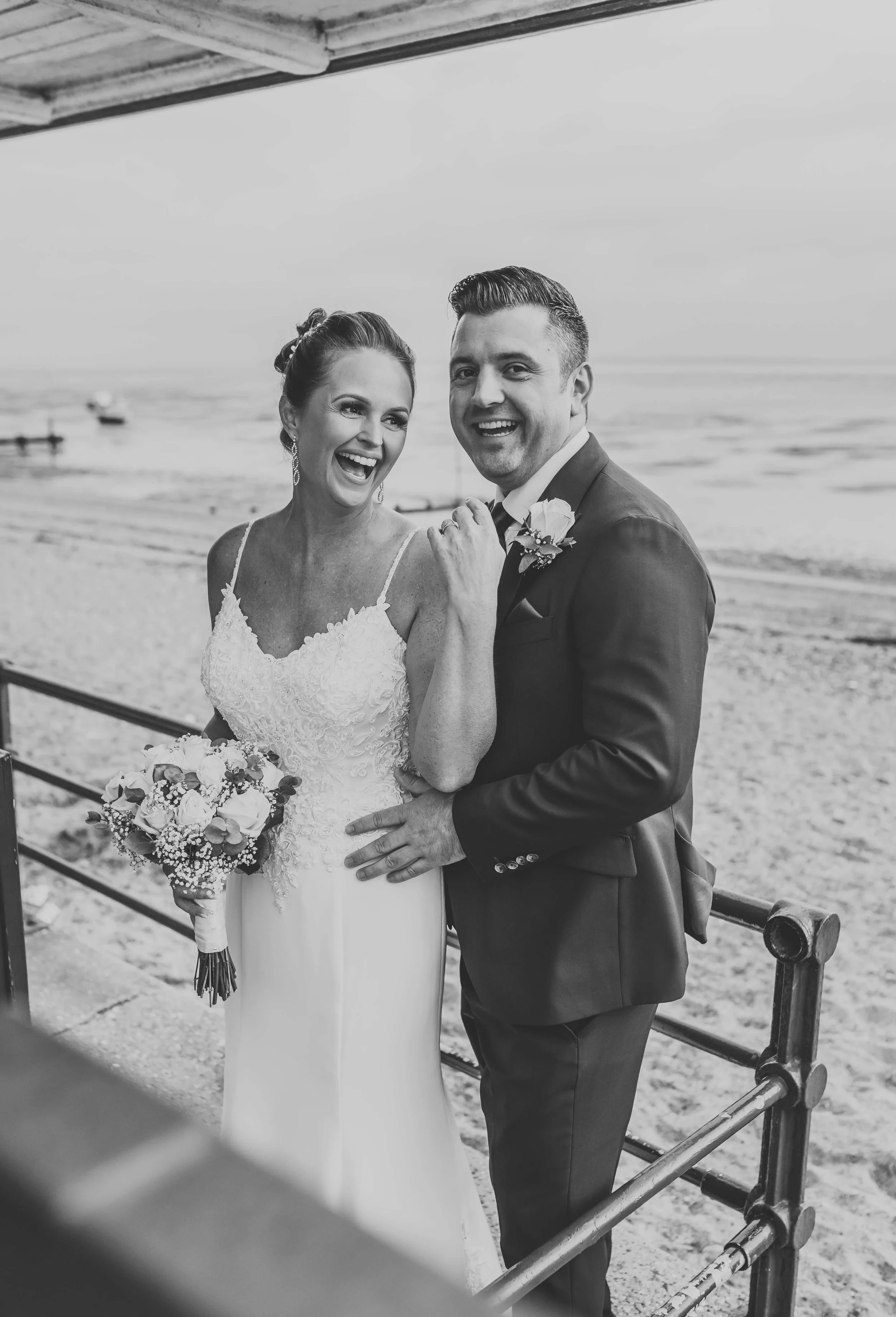 A black-and-white photo of a smiling bride and groom on the beach, celebrating their wedding. The bride is holding a bouquet and the groom is in a suit with a boutonniere. They appear happy and joyful.