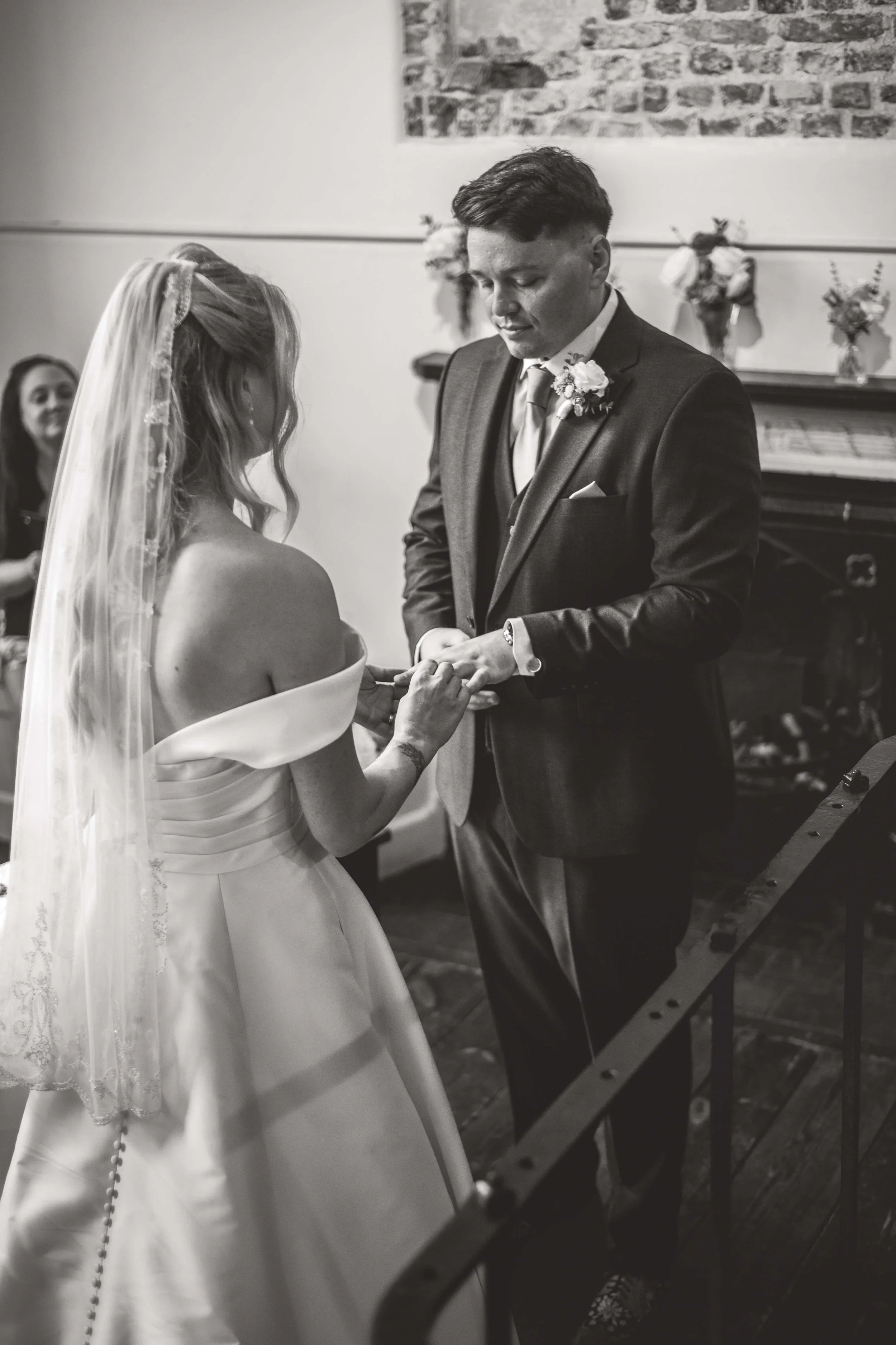 A black-and-white photo of a wedding ceremony showing a bride and groom exchanging rings. The bride is wearing an off-the-shoulder wedding dress and veil, and the groom is in a suit with a boutonniere. A woman in the background is smiling.
