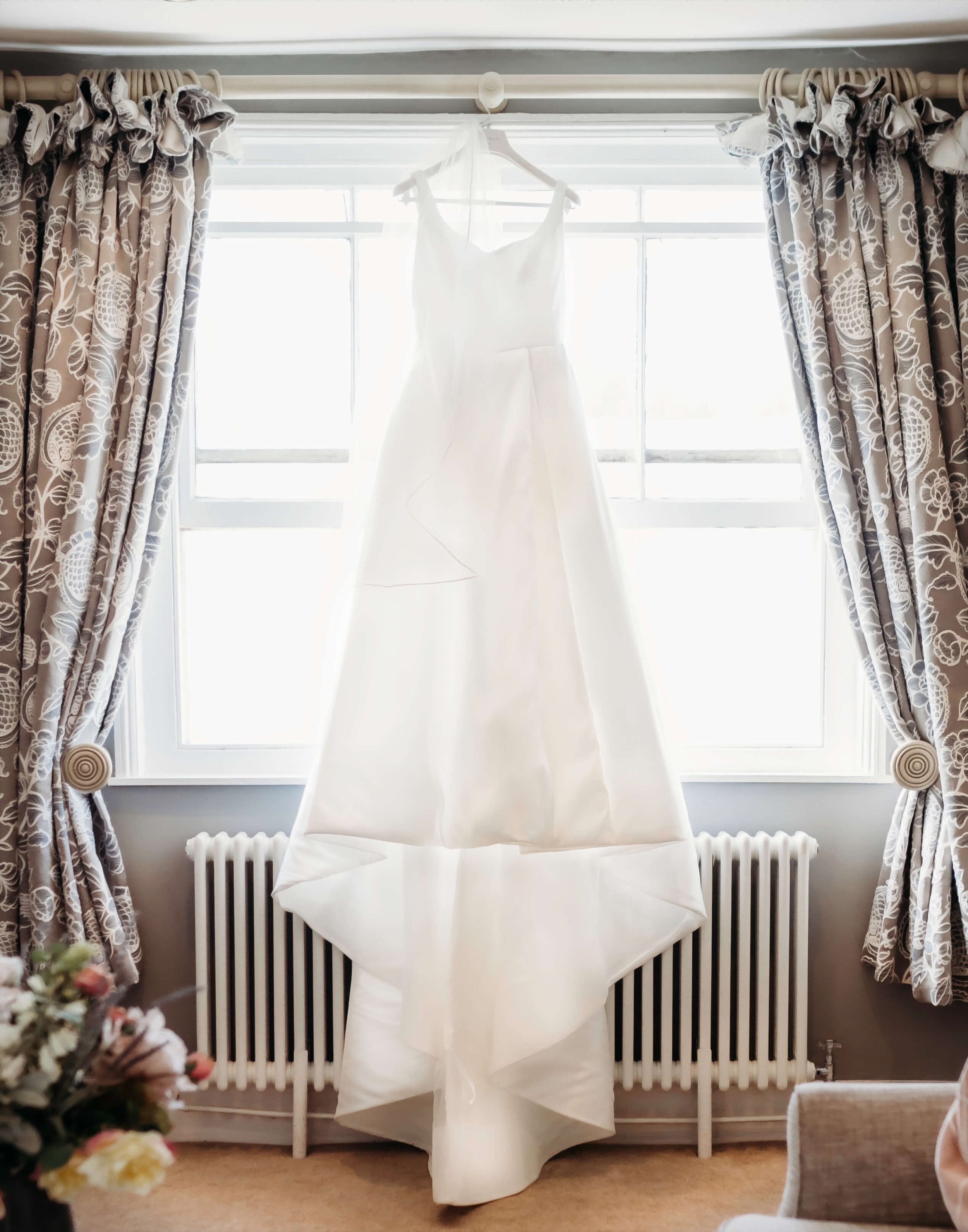 White wedding dress hanging in front of a window with patterned curtains on each side.