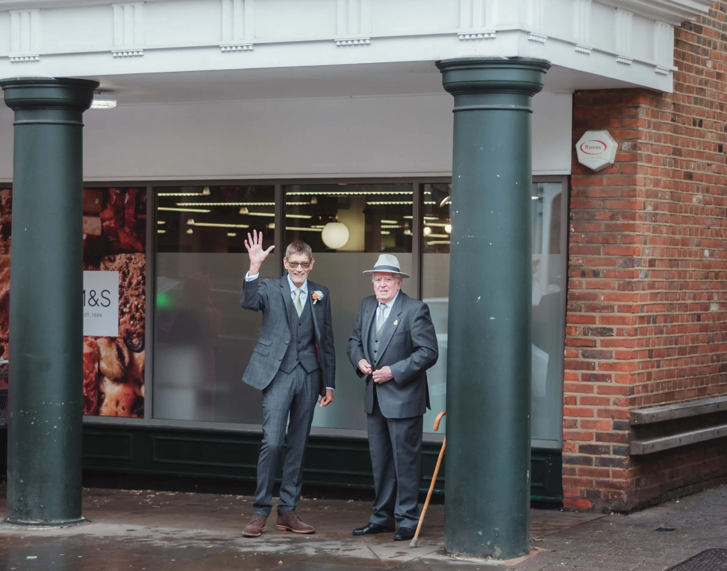 Two men in suits, one waving, standing on a sidewalk in front of a store window, with brick wall and green columns.