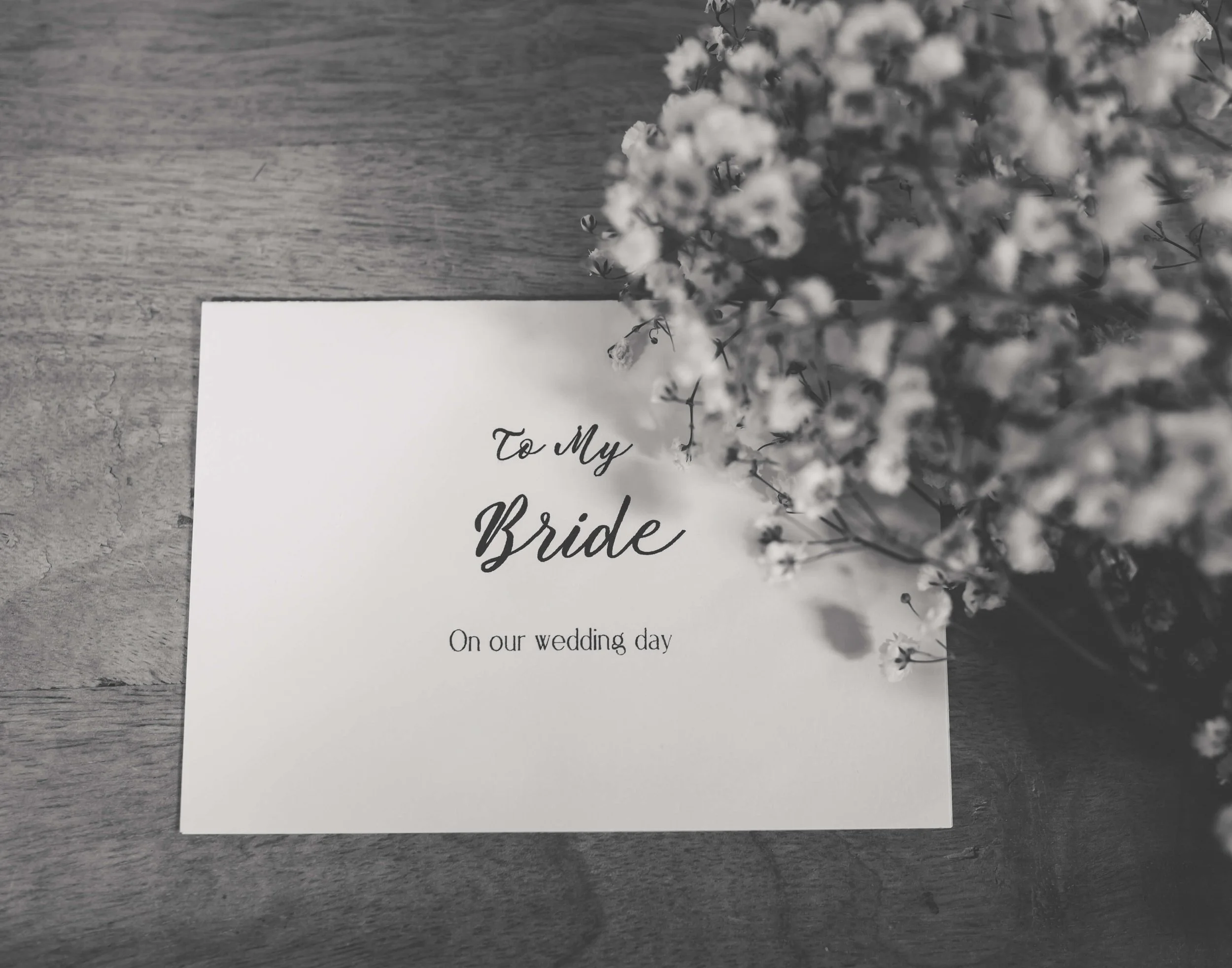 A wedding card on a wooden surface with a bouquet of small white flowers partially covering the top right corner.