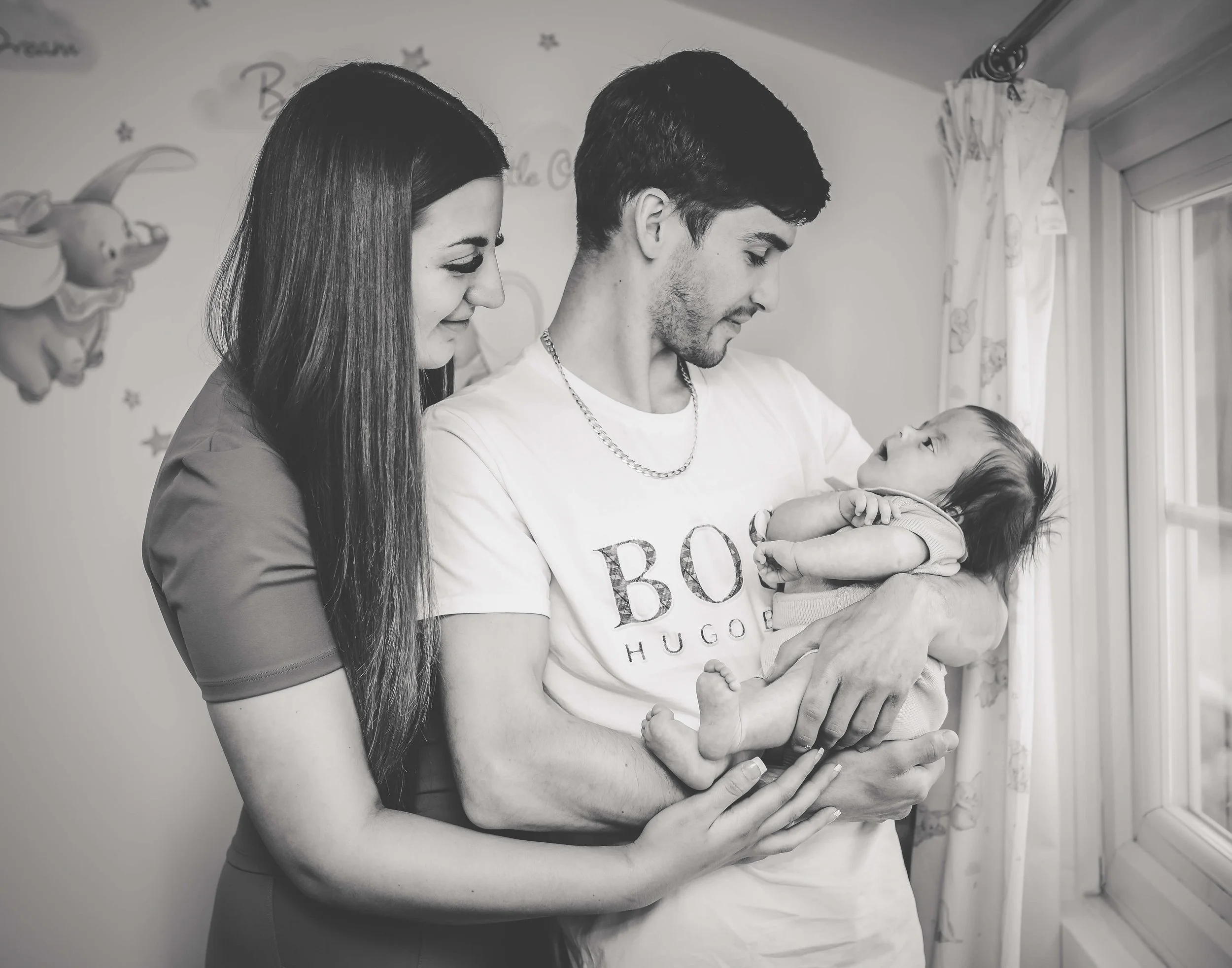 A family of three looking at a baby, with the father holding the baby and the mother touching the father's arm, inside a room with a window and curtains.