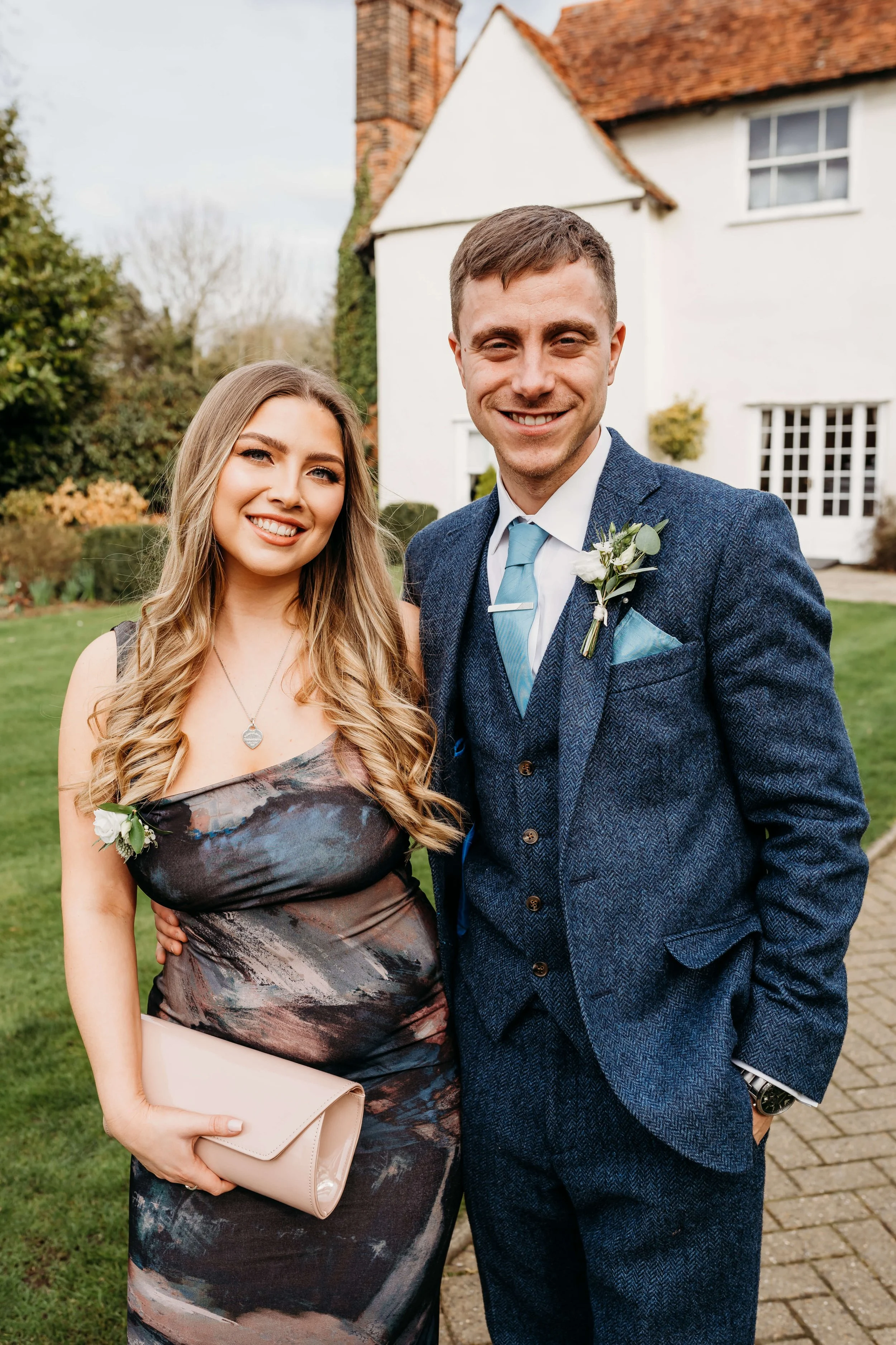 A young woman and a young man, dressed in formal attire, standing outdoors in front of a white house with a red-brick chimney, smiling at the camera.