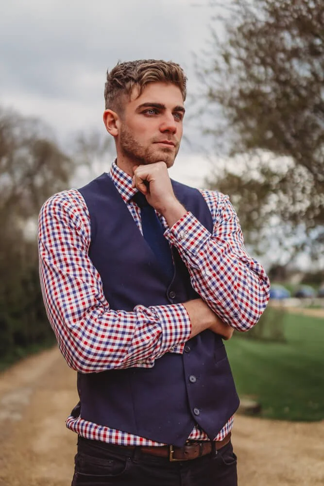 Young man with brown hair and beard, wearing a checkered shirt and navy vest, standing outdoors with trees and a cloudy sky in the background.