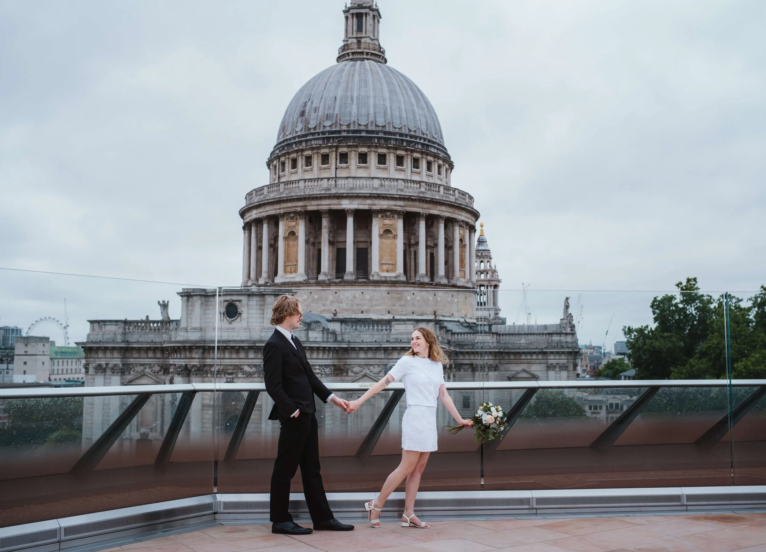 A couple holding hands on a rooftop with St. Paul's Cathedral in London in the background. The woman is holding a bouquet of flowers and wearing a white dress, while the man is in a black suit.