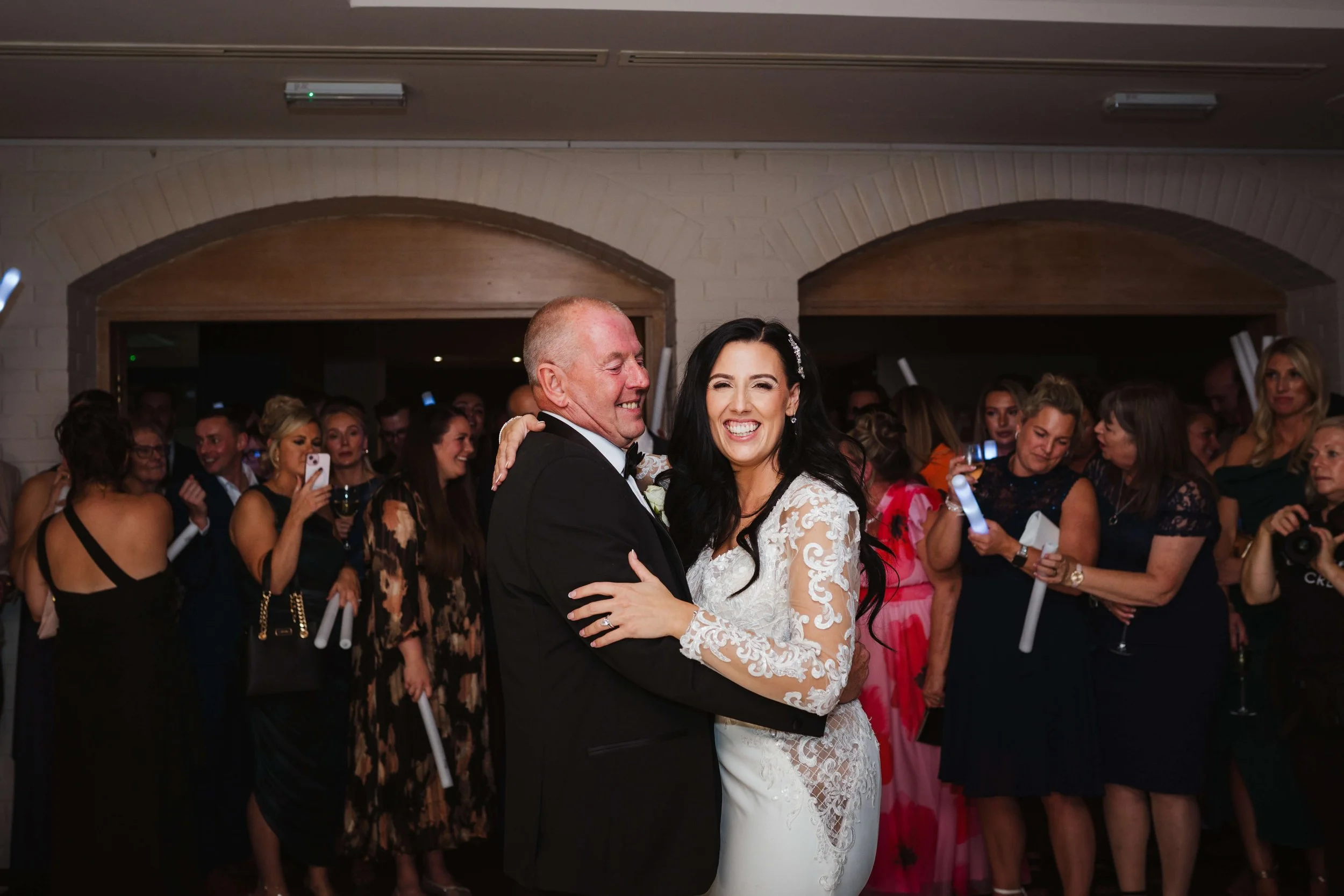 A bride and an older man, possibly her father, dancing and smiling at a wedding reception while guests watch and take photos in the background.