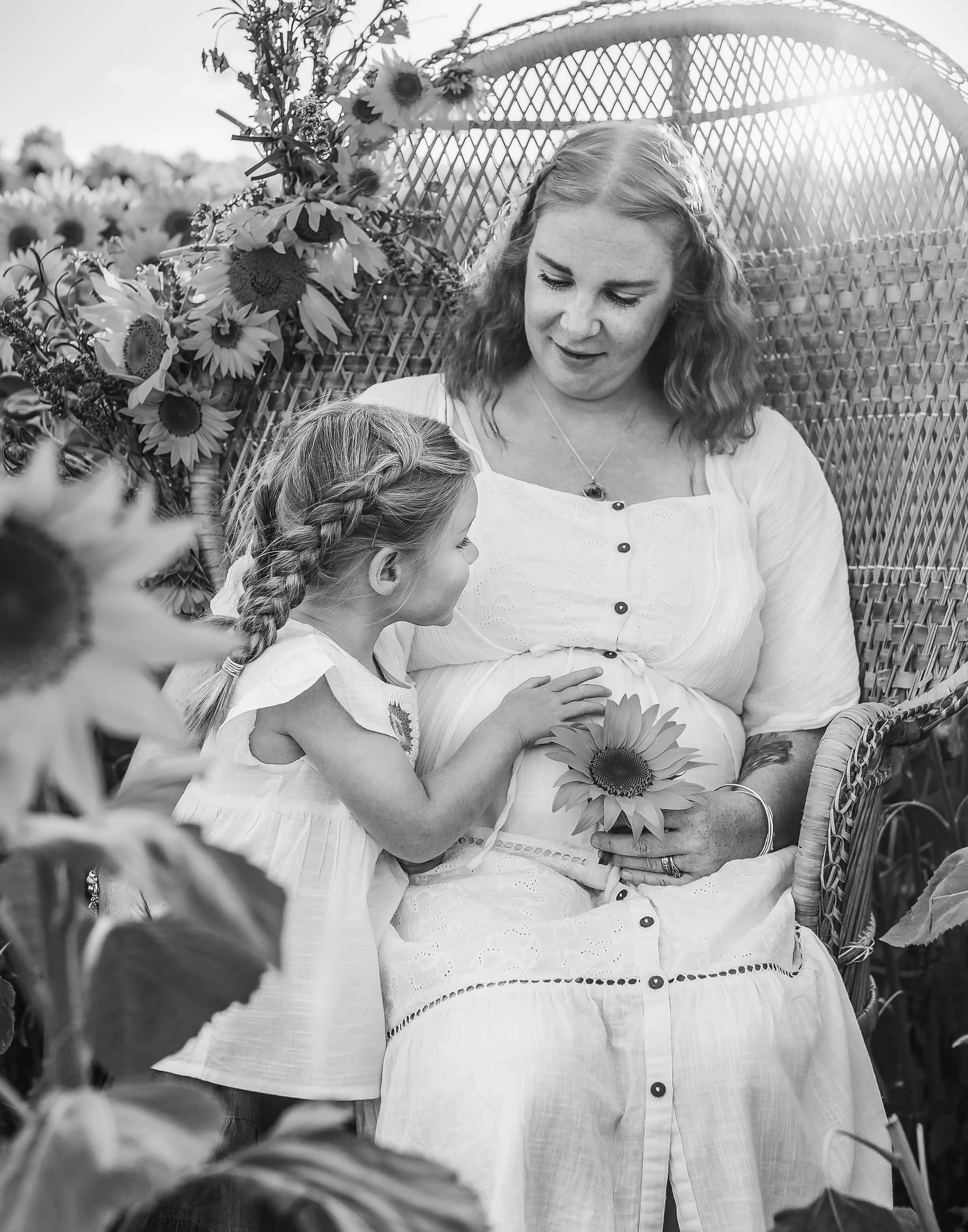 daughter touches mums pregnant belly with sun flare coming through the gaps in peacock chair 