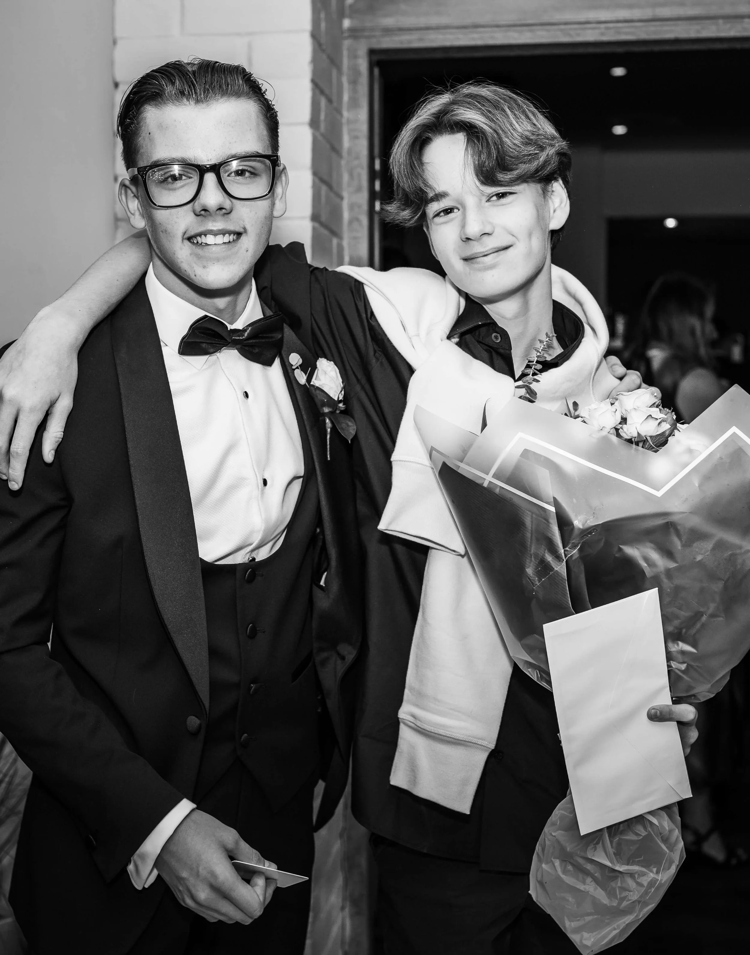 Two young men smiling, with one in a tuxedo and glasses and the other in casual clothing holding a bouquet of flowers.