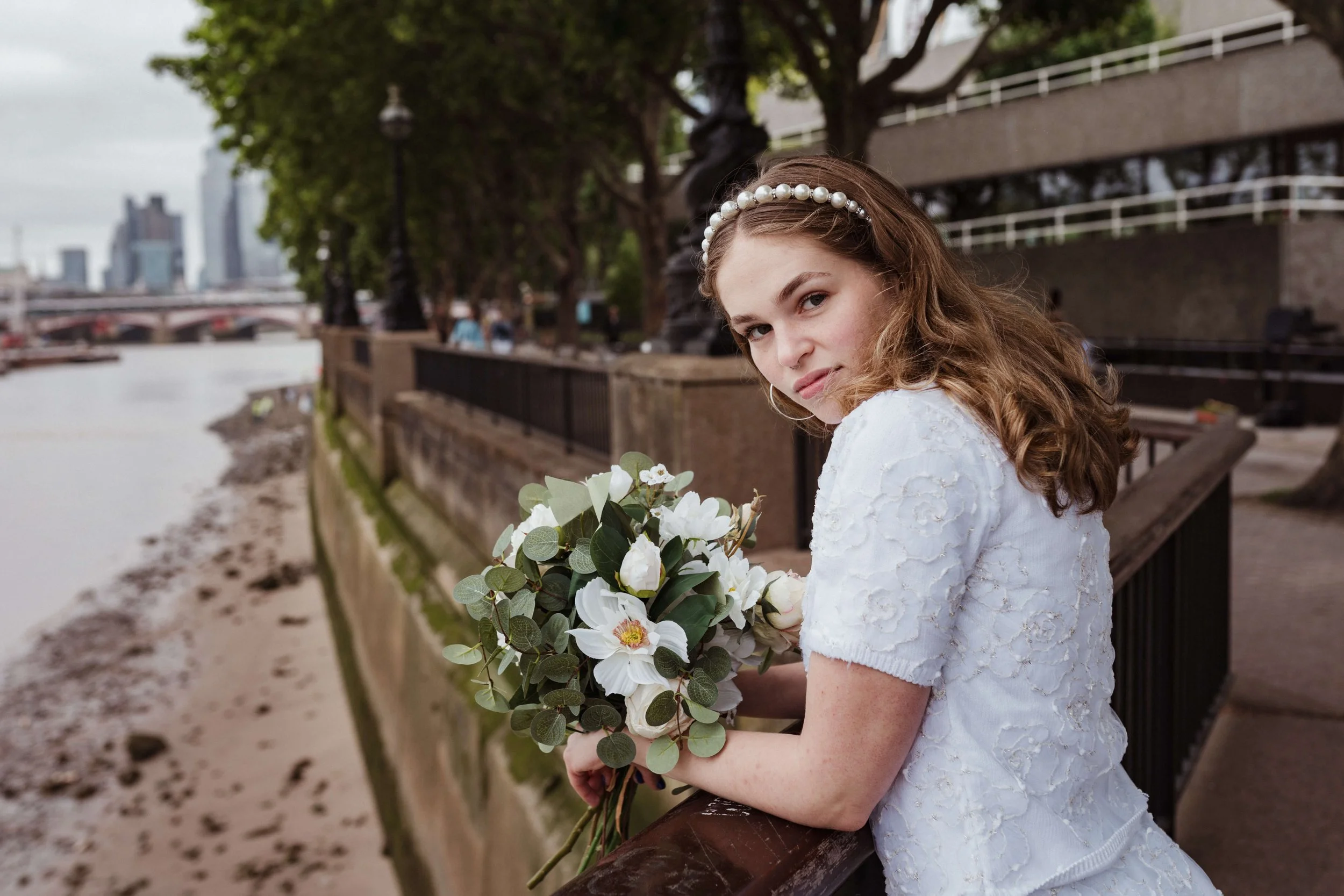 Young girl in white dress wearing pearl headband holding a bouquet of white flowers on a riverside promenade with trees and city skyline in the background.
