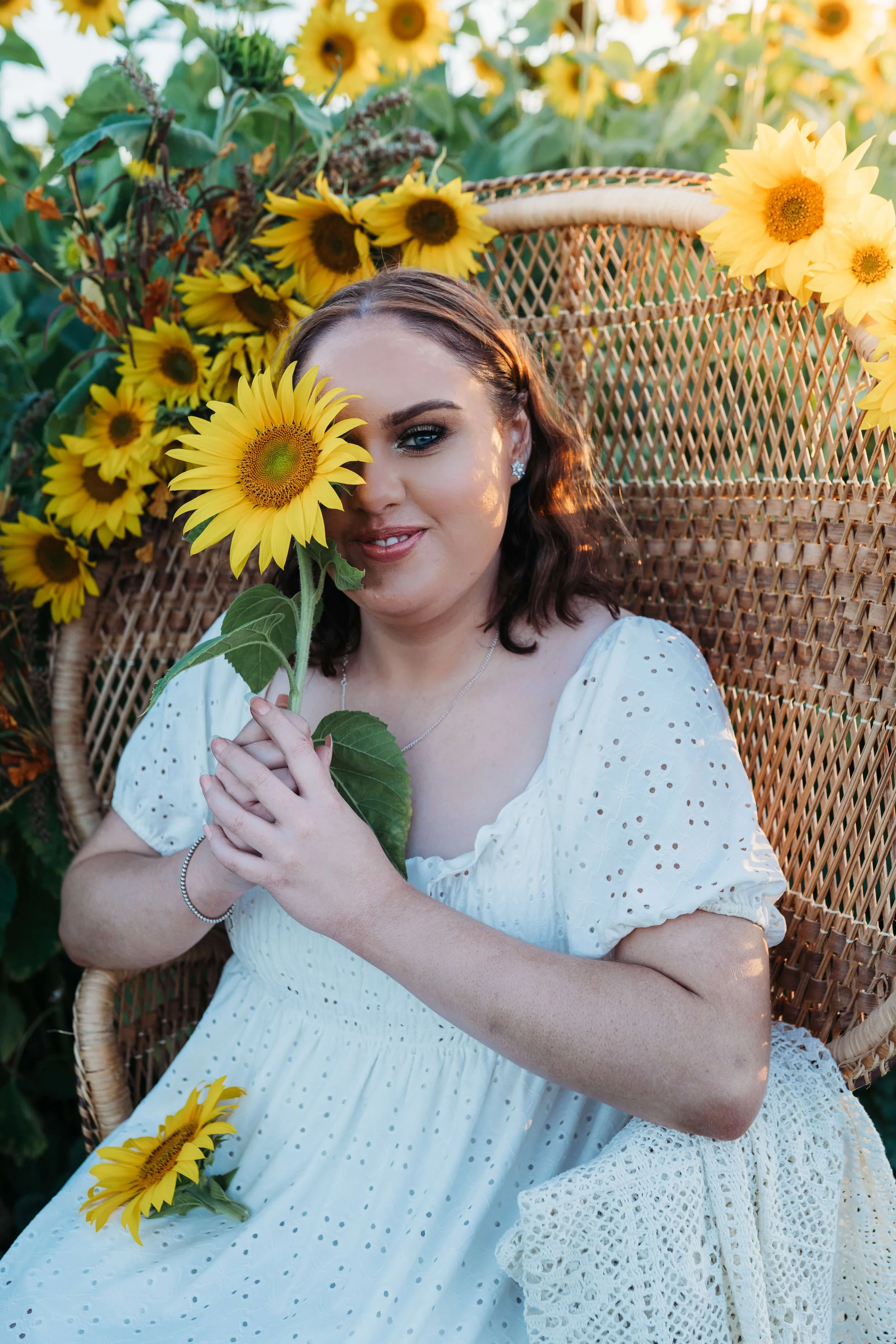 A woman with dark brown hair and blue eyes sitting on a woven chair outdoors, holding a sunflower to her face, surrounded by sunflowers in a field, wearing a white eyelet dress, with sunlight casting warm light on her.