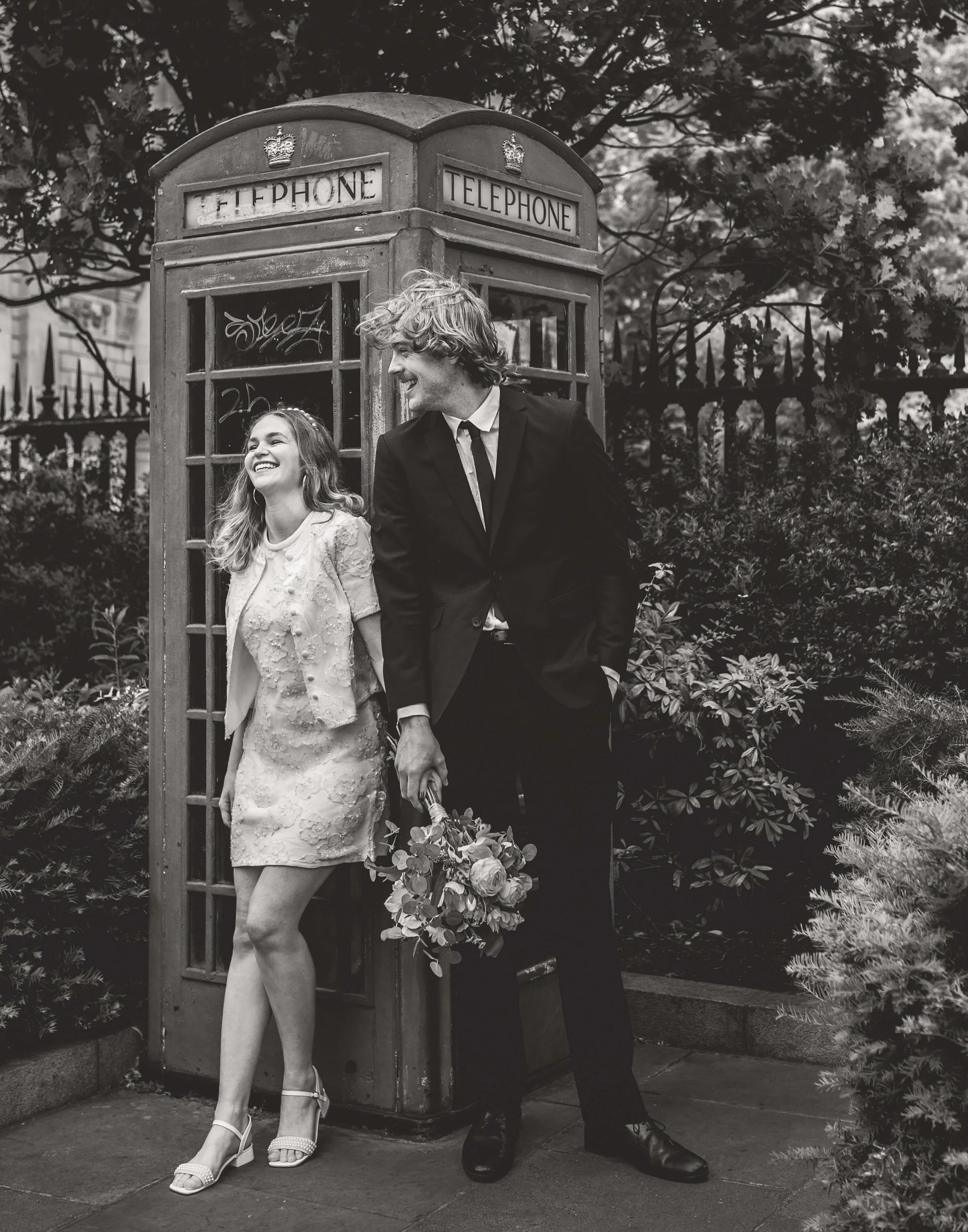 A couple, a woman in a dress and heels and a man in a suit, standing next to a vintage telephone booth outdoors, smiling and laughing, with the woman holding a bouquet of flowers.