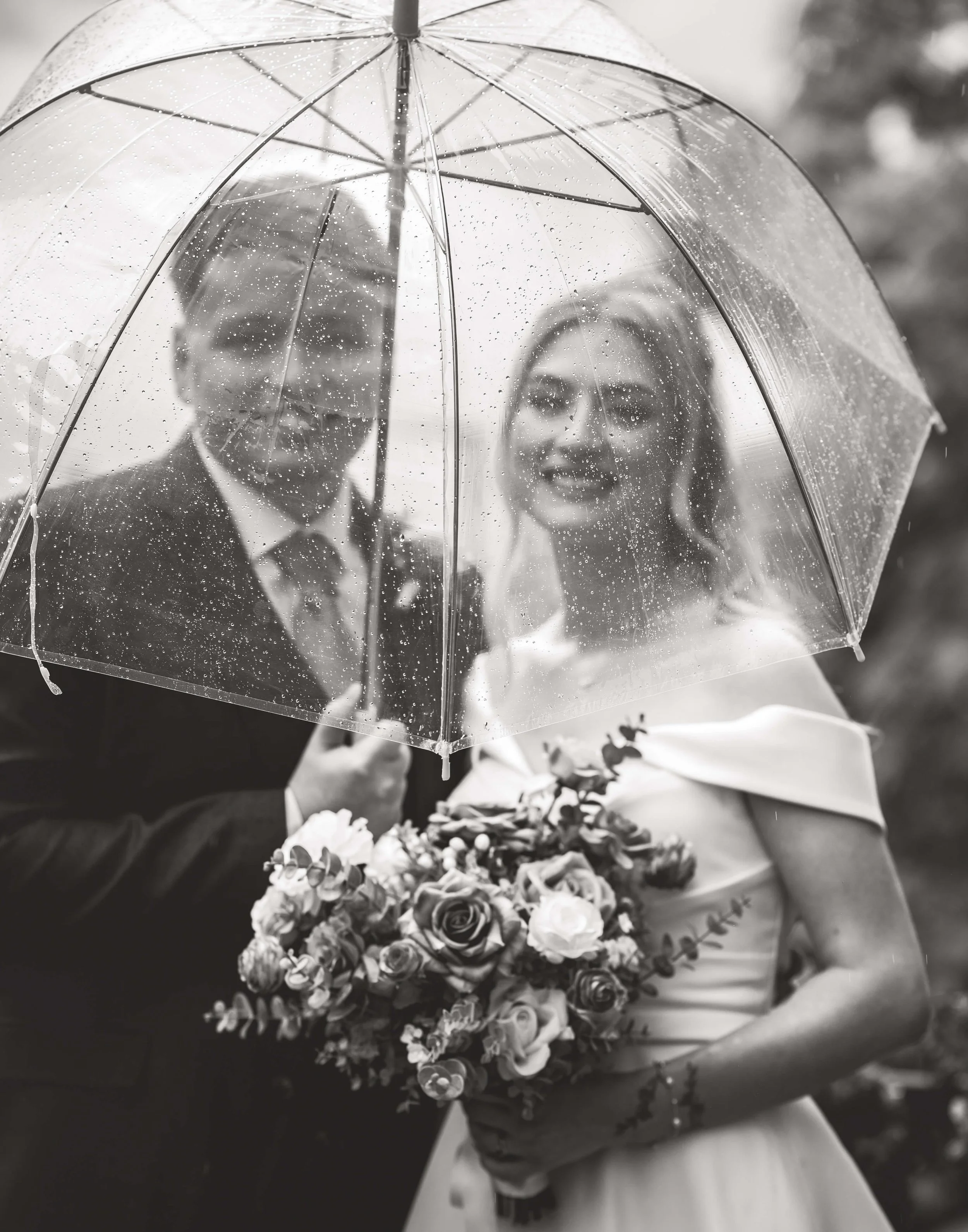 A black and white photo of a bride and groom standing close under a transparent umbrella with rain droplets on it. The bride is holding a bouquet of flowers and smiling, and the groom is dressed in a suit with a tie, also smiling.