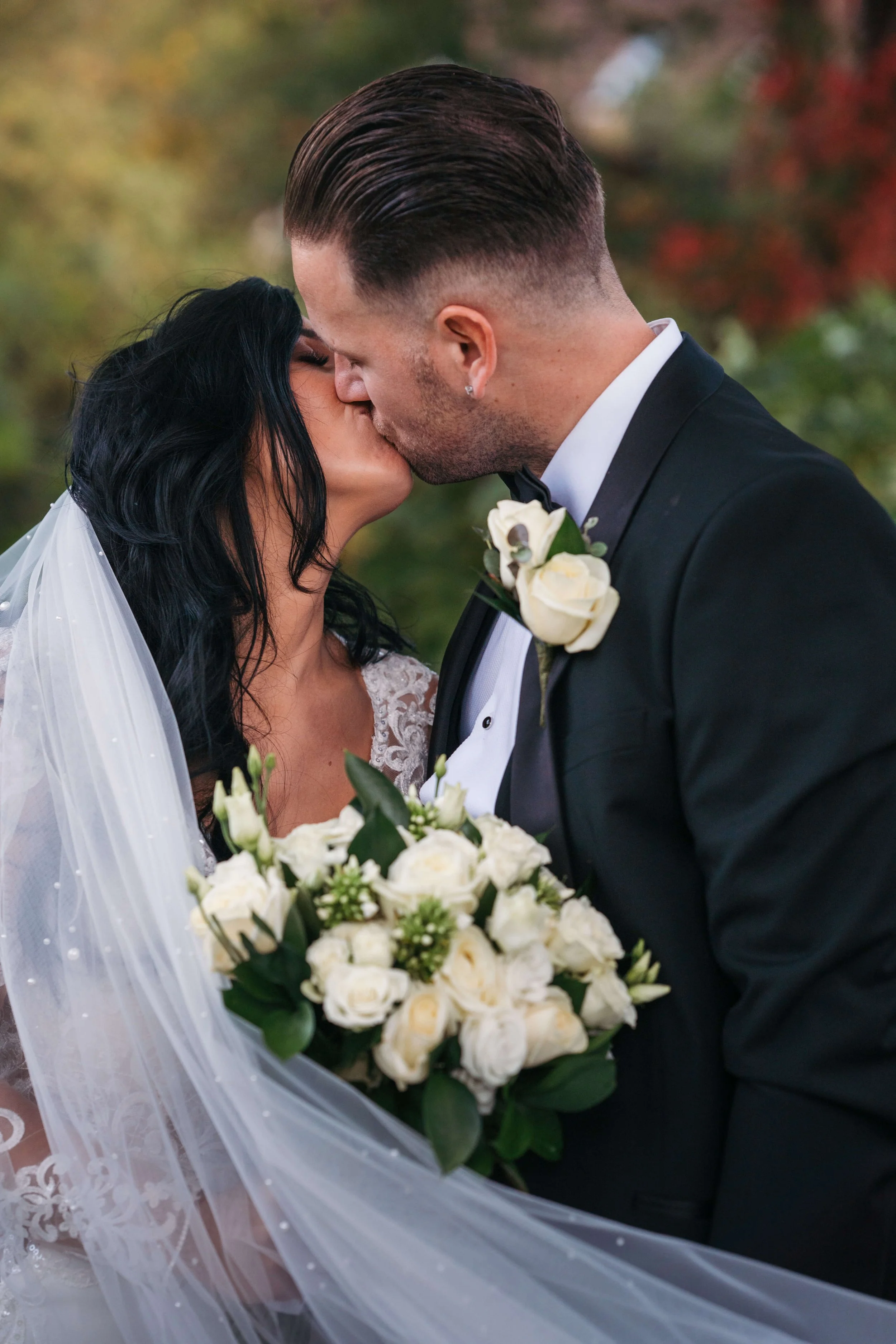 A bride and groom kissing outdoors during a wedding, with the bride holding a bouquet of white roses and wearing a veil and lace wedding dress, while the groom wears a black tuxedo with a white shirt, black bow tie, and a white rose boutonniere.