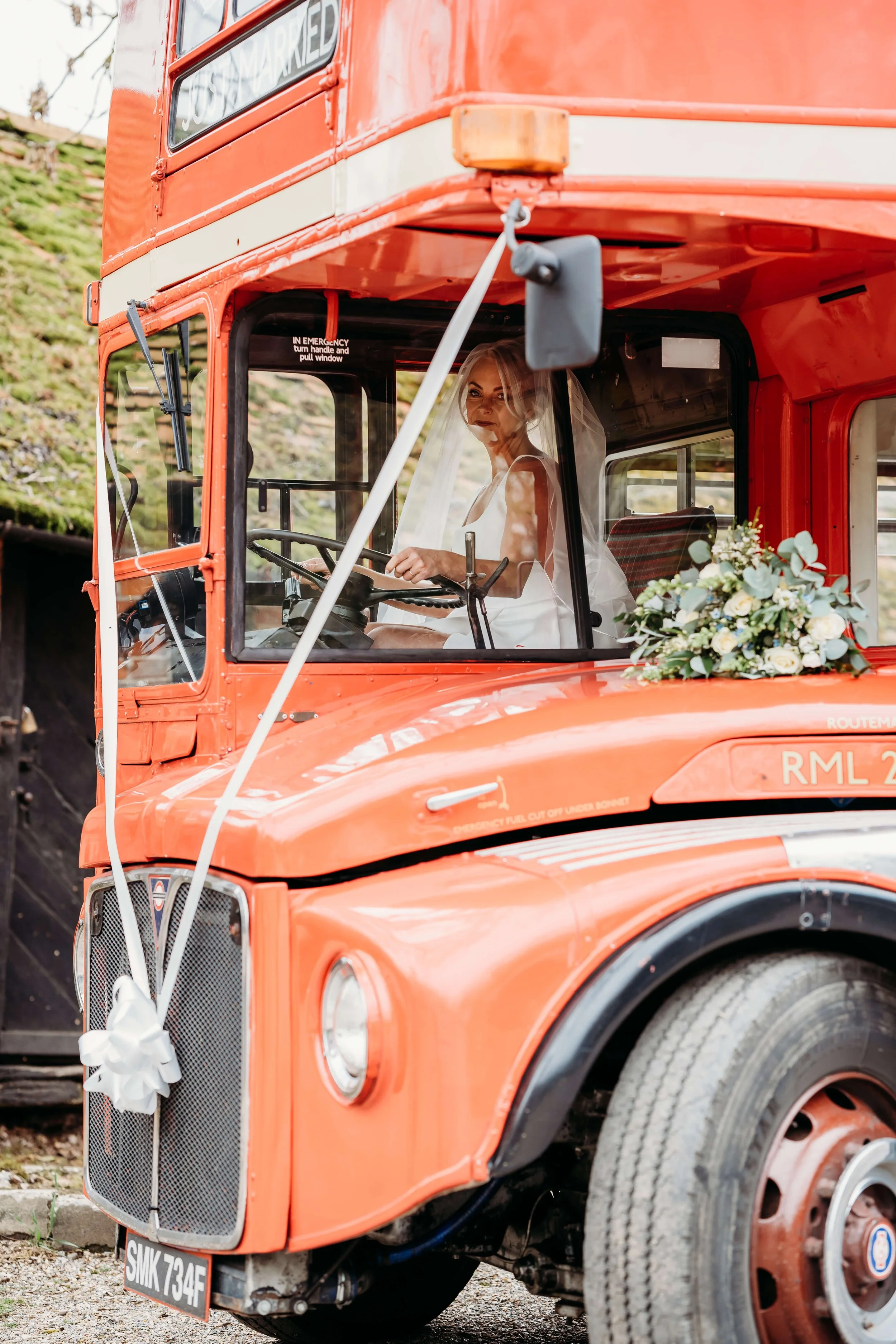 A bride wearing a white wedding dress and veil sitting inside a vintage orange double-decker bus decorated with white flowers and a white ribbon bow. The bride is looking out the window.
