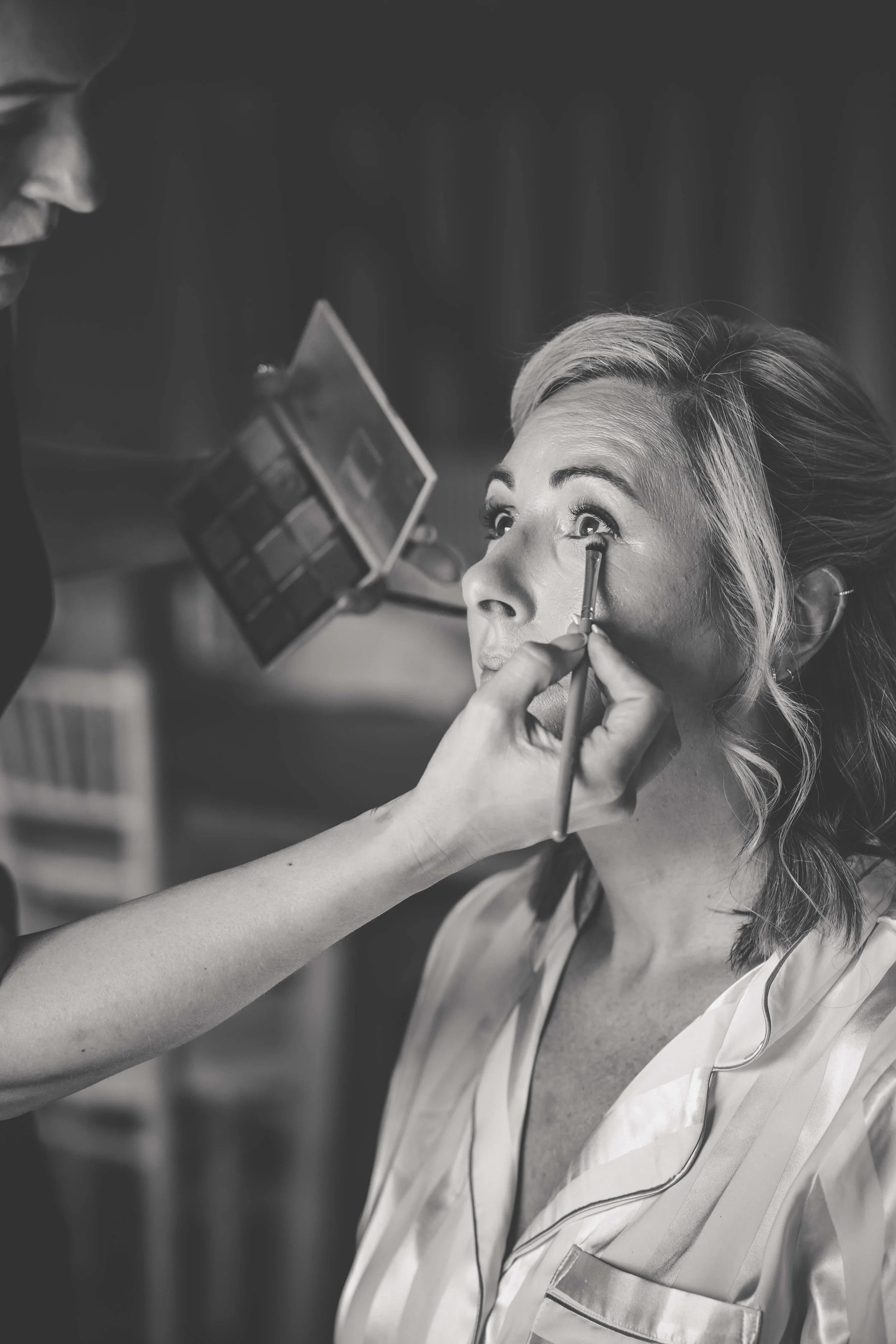 Makeup artist applying eye makeup to a woman with wavy hair in a salon or studio.