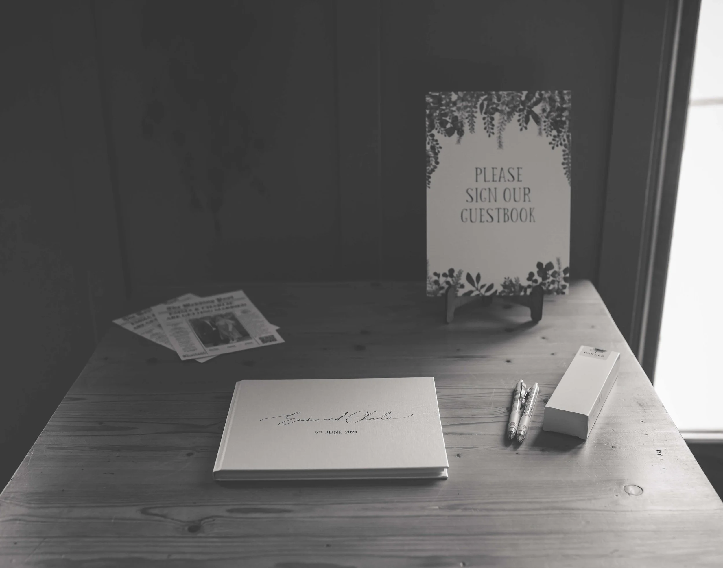 Guestbook with floral design on a table, along with pens and a box, near a sign that says "Please Sign Our Guestbook".