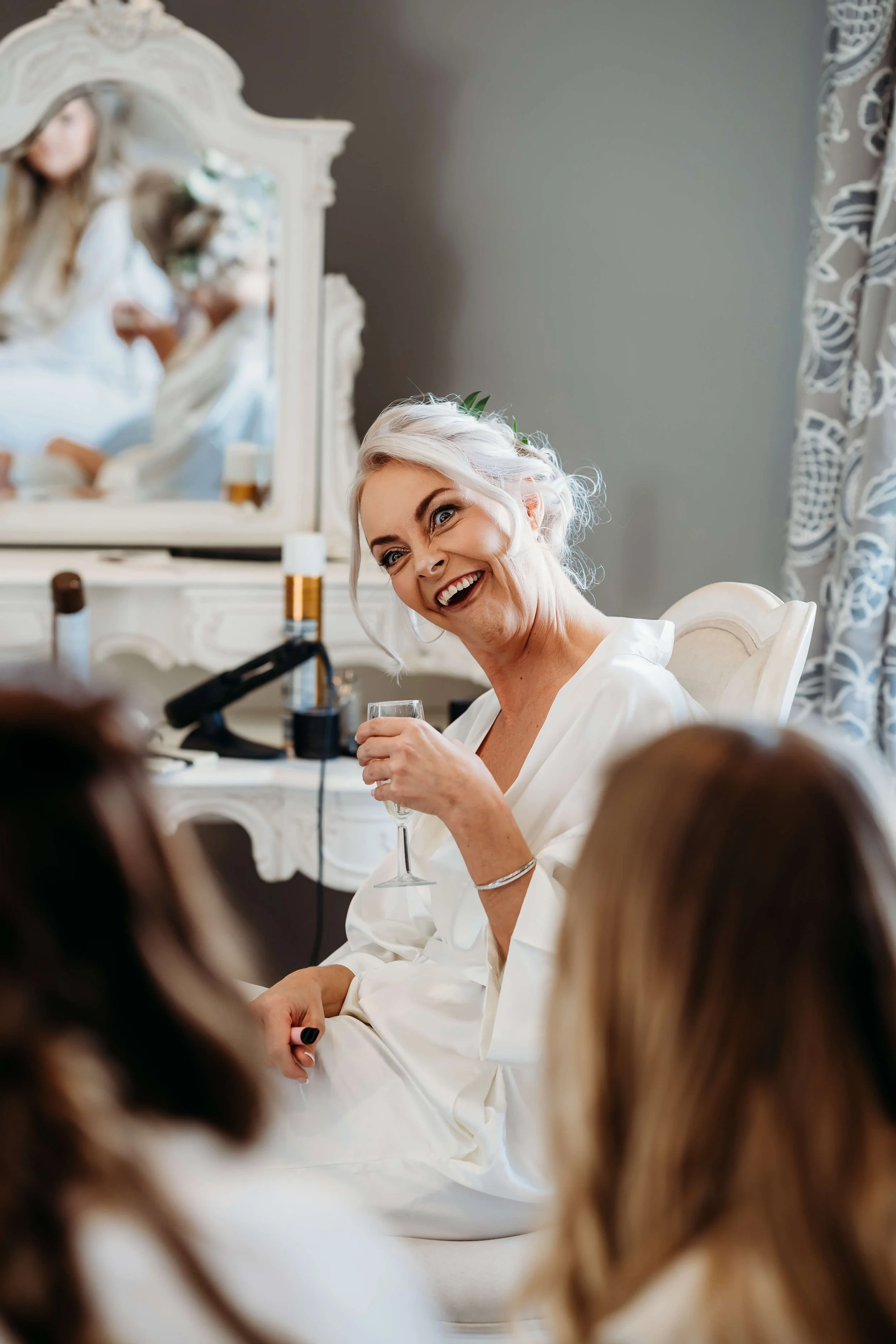 A joyful woman with platinum blonde hair styled in loose waves, wearing a white robe, sitting in a decorated room with a vintage dresser and mirror reflected in the background, holding a glass of wine and smiling at someone in front of her.