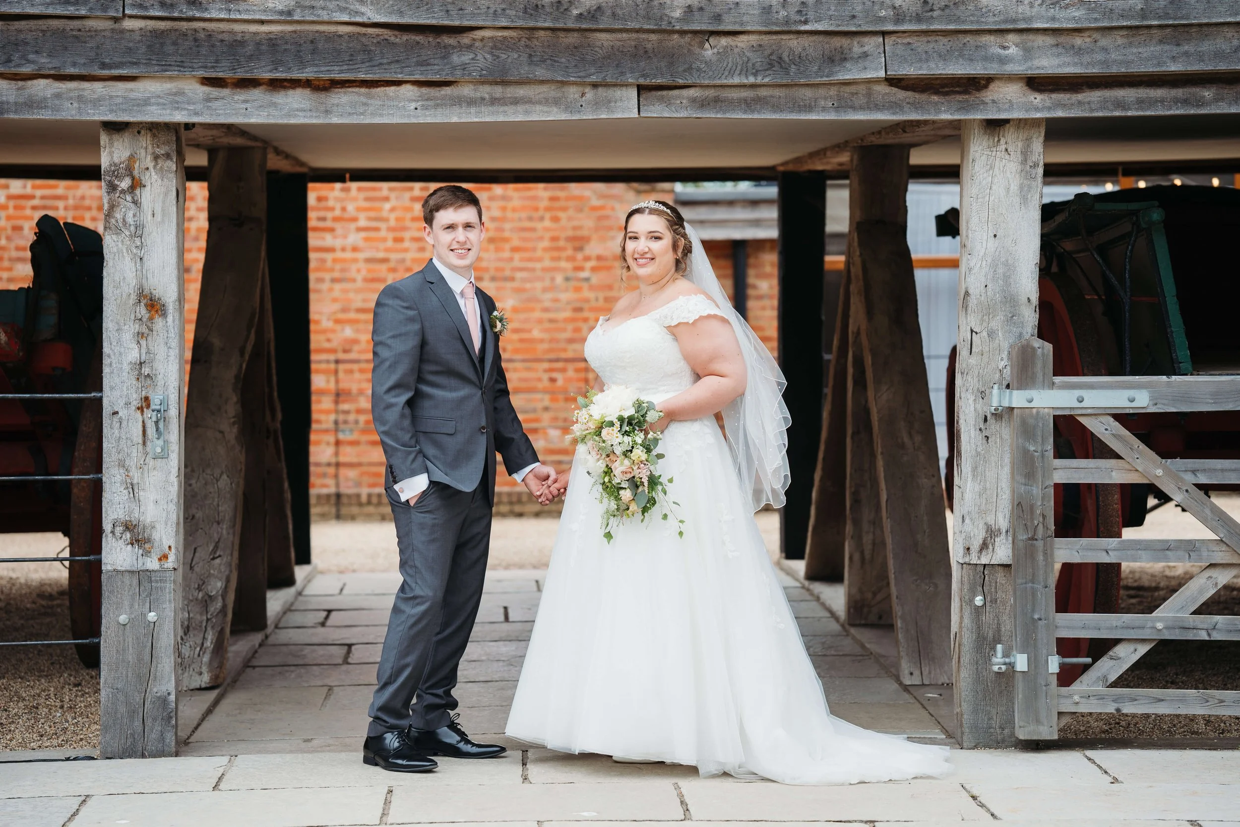 A bride and groom standing hand in hand under a rustic wooden structure outdoors, with brick wall and farm equipment in the background. The bride is in a white wedding dress holding a bouquet, and the groom is in a gray suit with a pink tie.