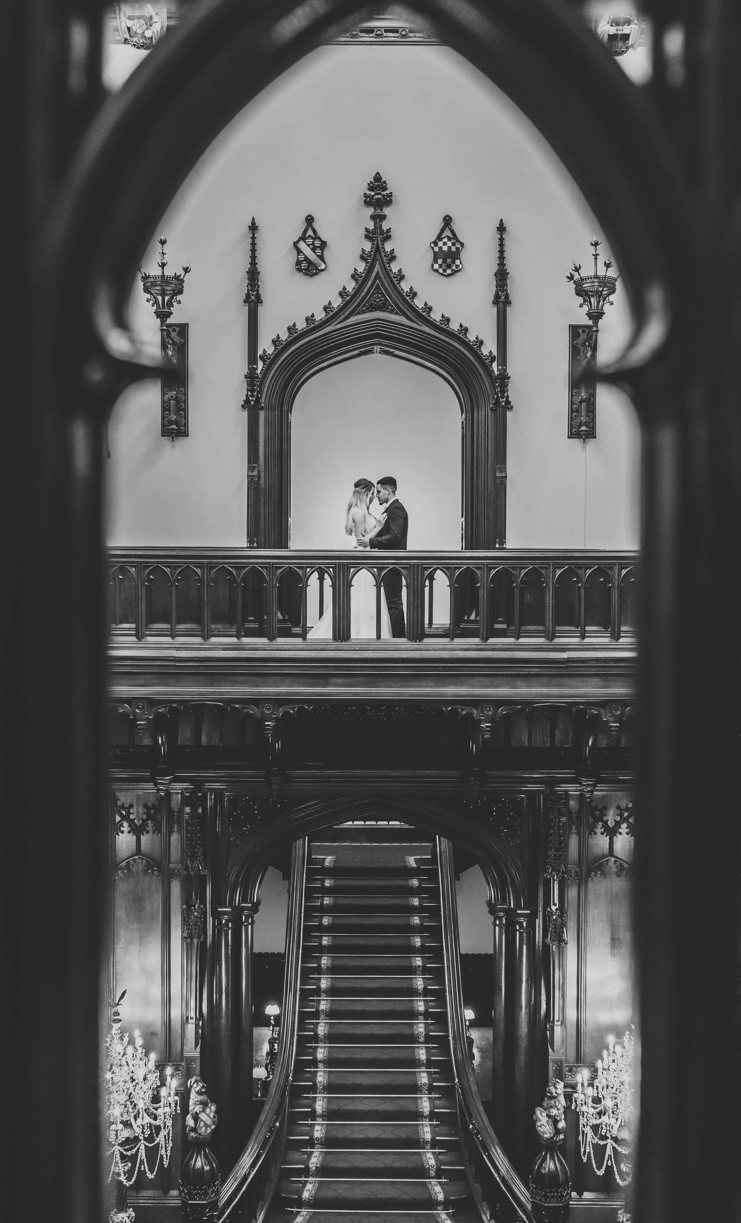 Black and white photo of a bride and groom embracing on a grand, ornate staircase with gothic architectural features, including decorative arches and crest emblems.