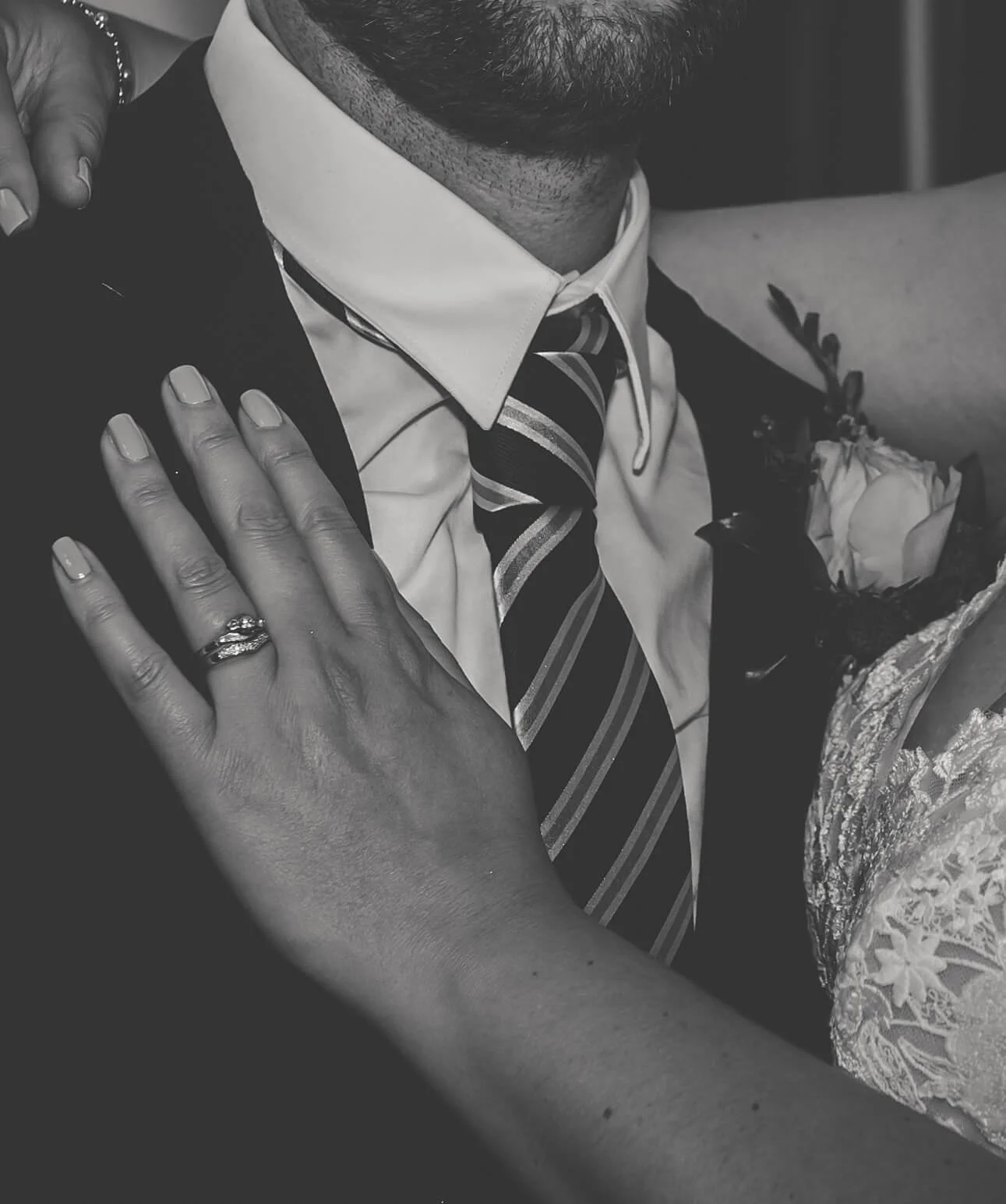 Close-up of a man in a suit with a tie, showing a woman’s hand with a wedding ring resting on his chest, and a woman in a dress with lace details hugging him with a corsage on her wrist.