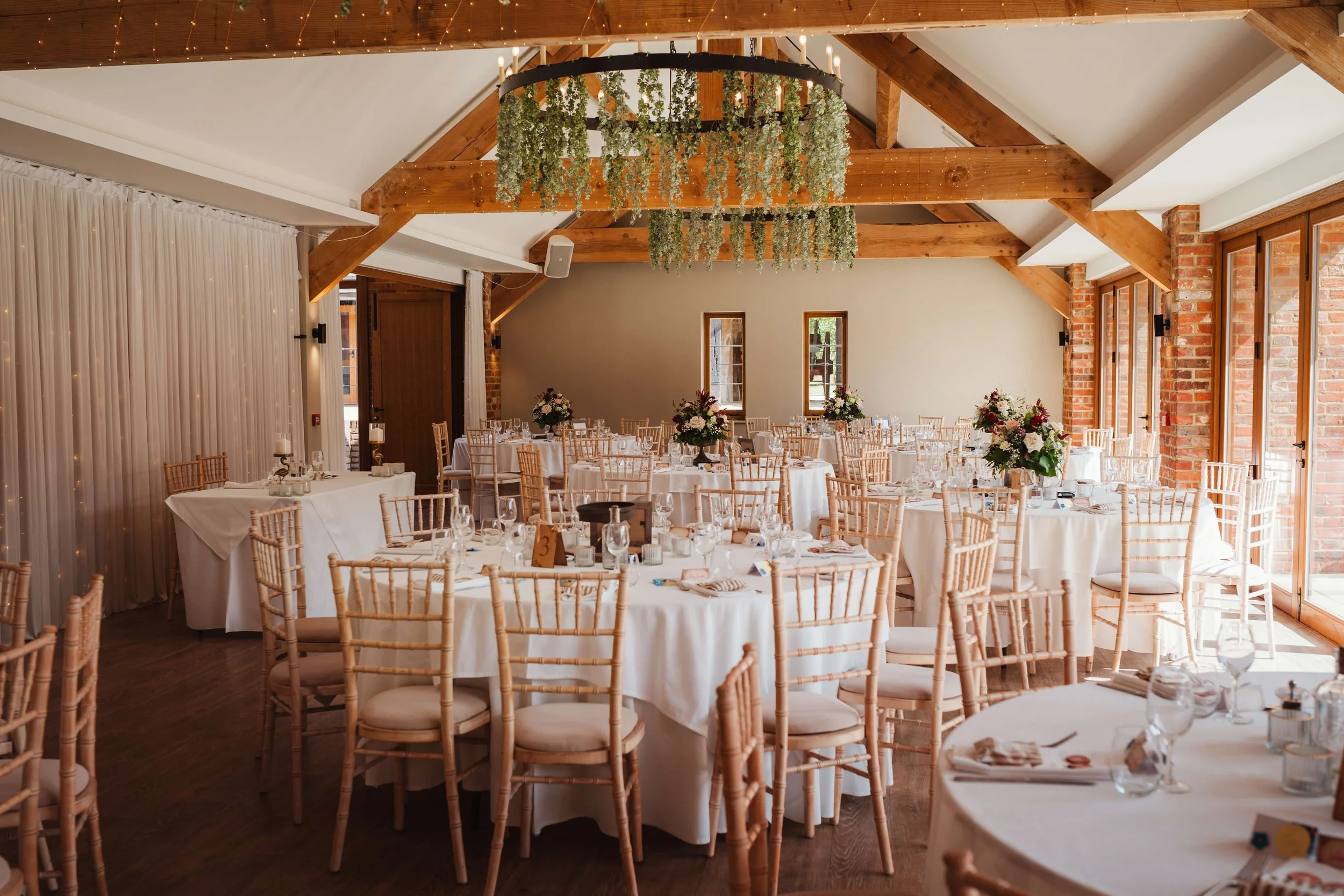 Elegant banquet hall decorated for a wedding reception with round tables draped in white cloth, surrounded by beige chairs, flowers centerpieces, and large windows allowing natural light, with wooden beams and greenery hanging from the ceiling.