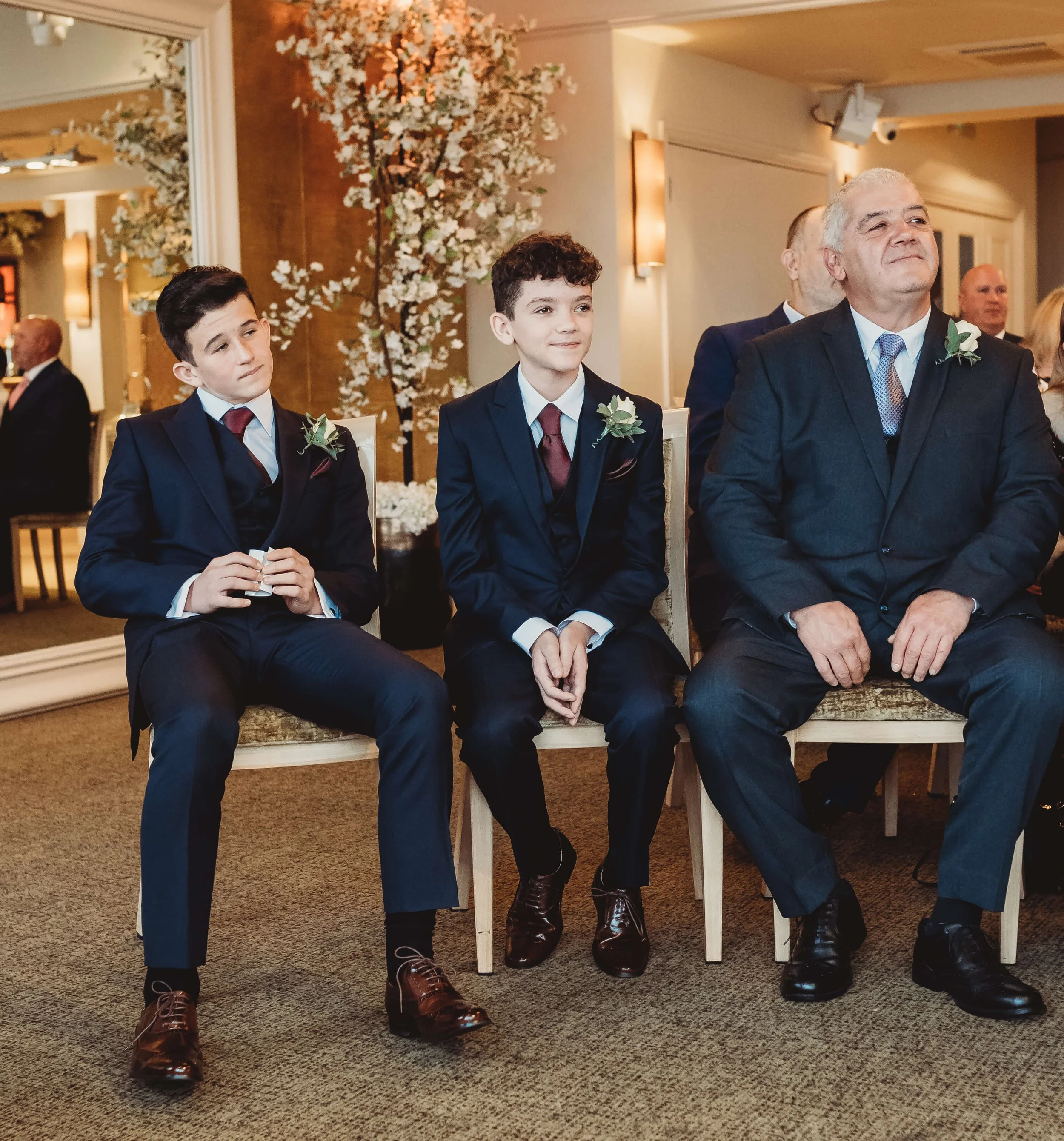 Three males in formal suits sitting in a row at a wedding or formal event, with floral decorations and background guests.